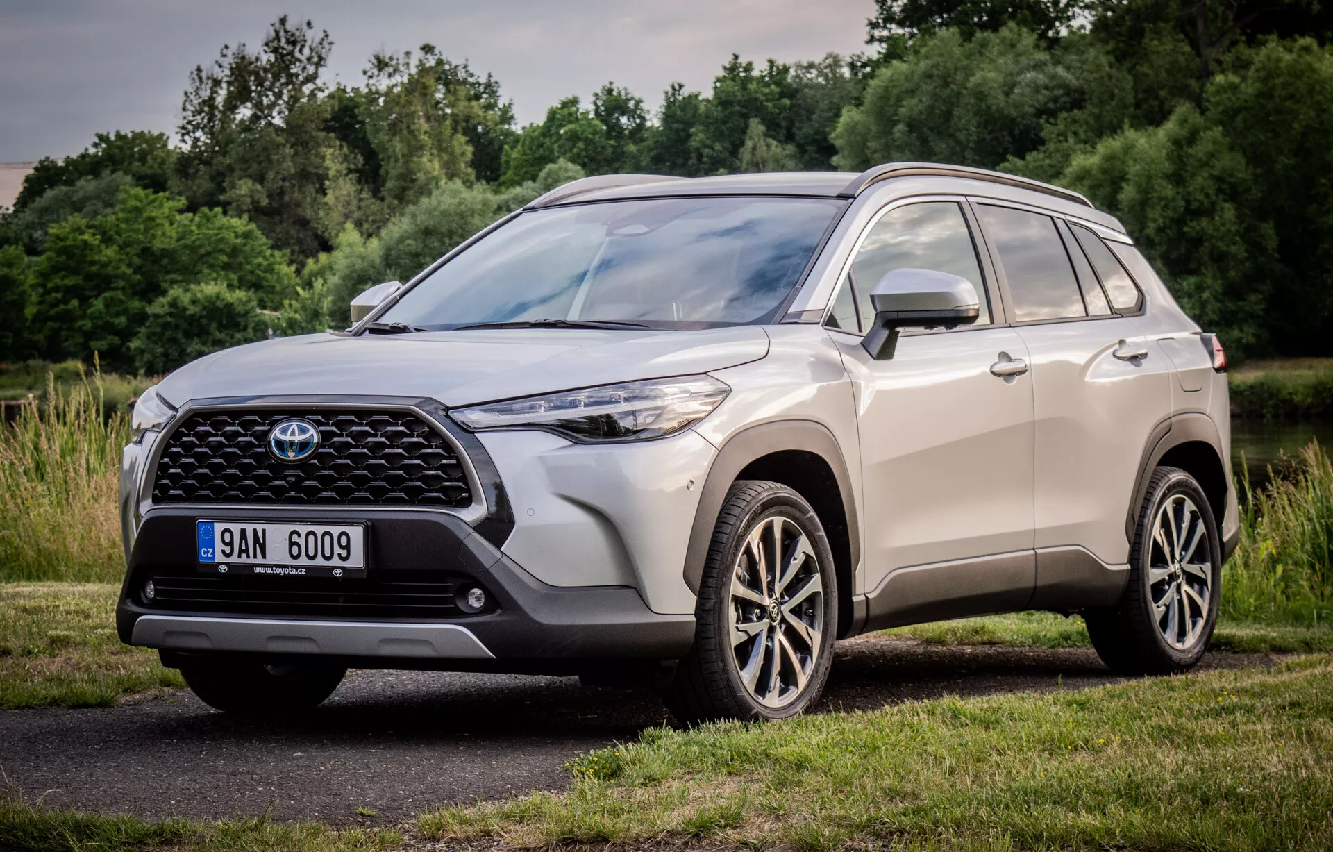 Toyota Corolla Cross 2022 che mostra il suo design dinamico con una vista in prospettiva frontale e laterale di tre quarti, evidenziando la grande griglia frontale sagomata, unità fari a LED eleganti e ruote in lega a più razze, parcheggiata in un ambiente naturale