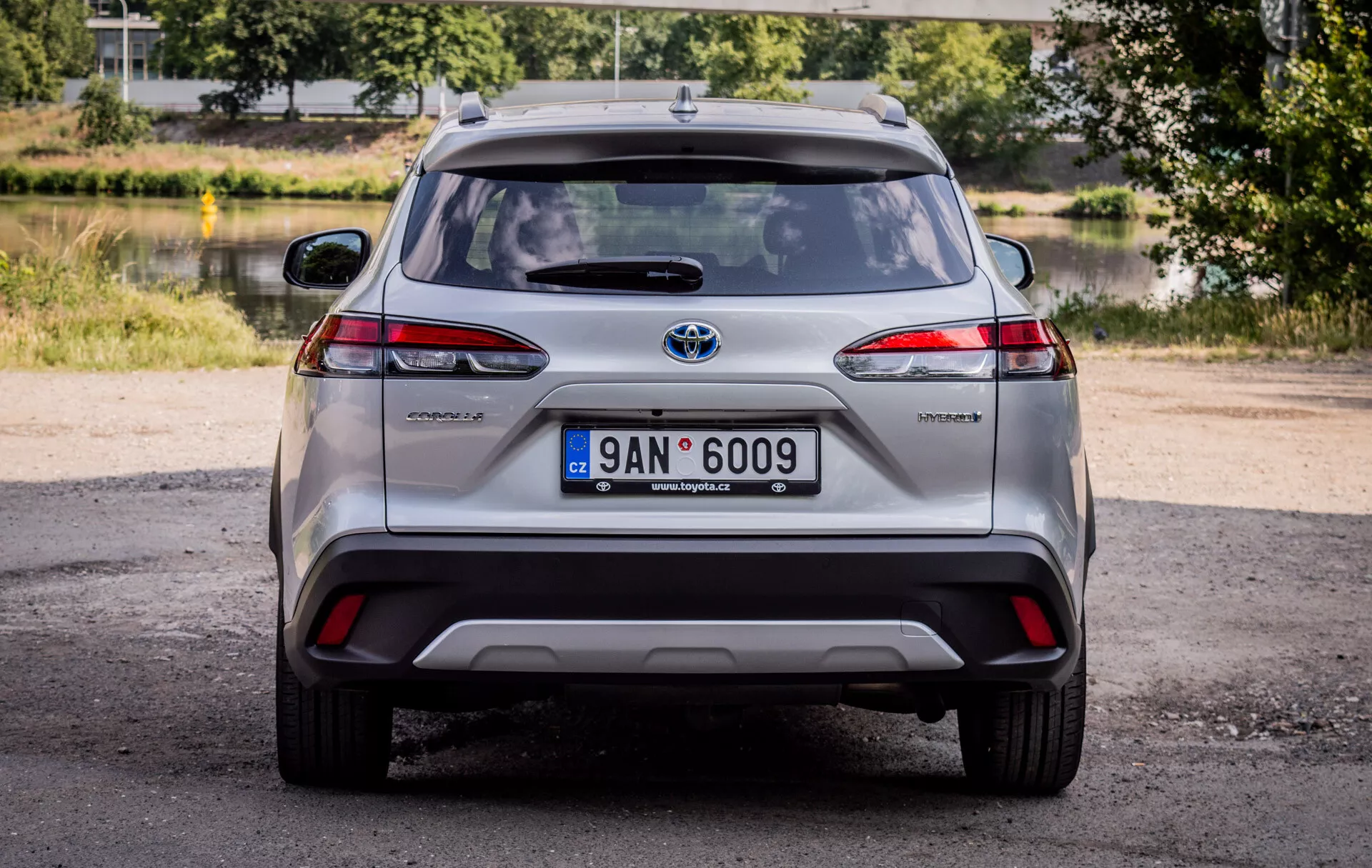 Vista posteriore della Toyota Corolla Cross 2022, con fari posteriori LED, badge ibrido e emblema Toyota cromato in un'ambientazione naturale diurna e uno sfondo scenico di verde e acqua