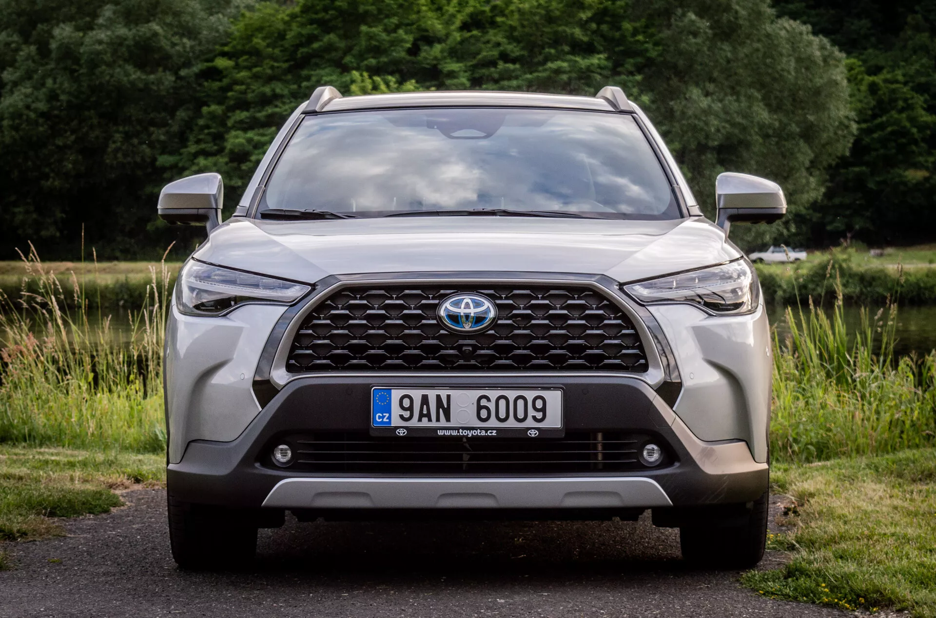 Vista frontale di una Toyota Corolla Cross 2022, che mette in risalto la sua grande griglia con emblema, fari moderni a LED, luci antinebbia, posizione della targa, sensori di parcheggio integrati o telecamera frontale, contorni aerodinamici del cofano, design robusto del paraurti inferiore e prese d'aria, parcheggiata su asfalto con uno sfondo naturale