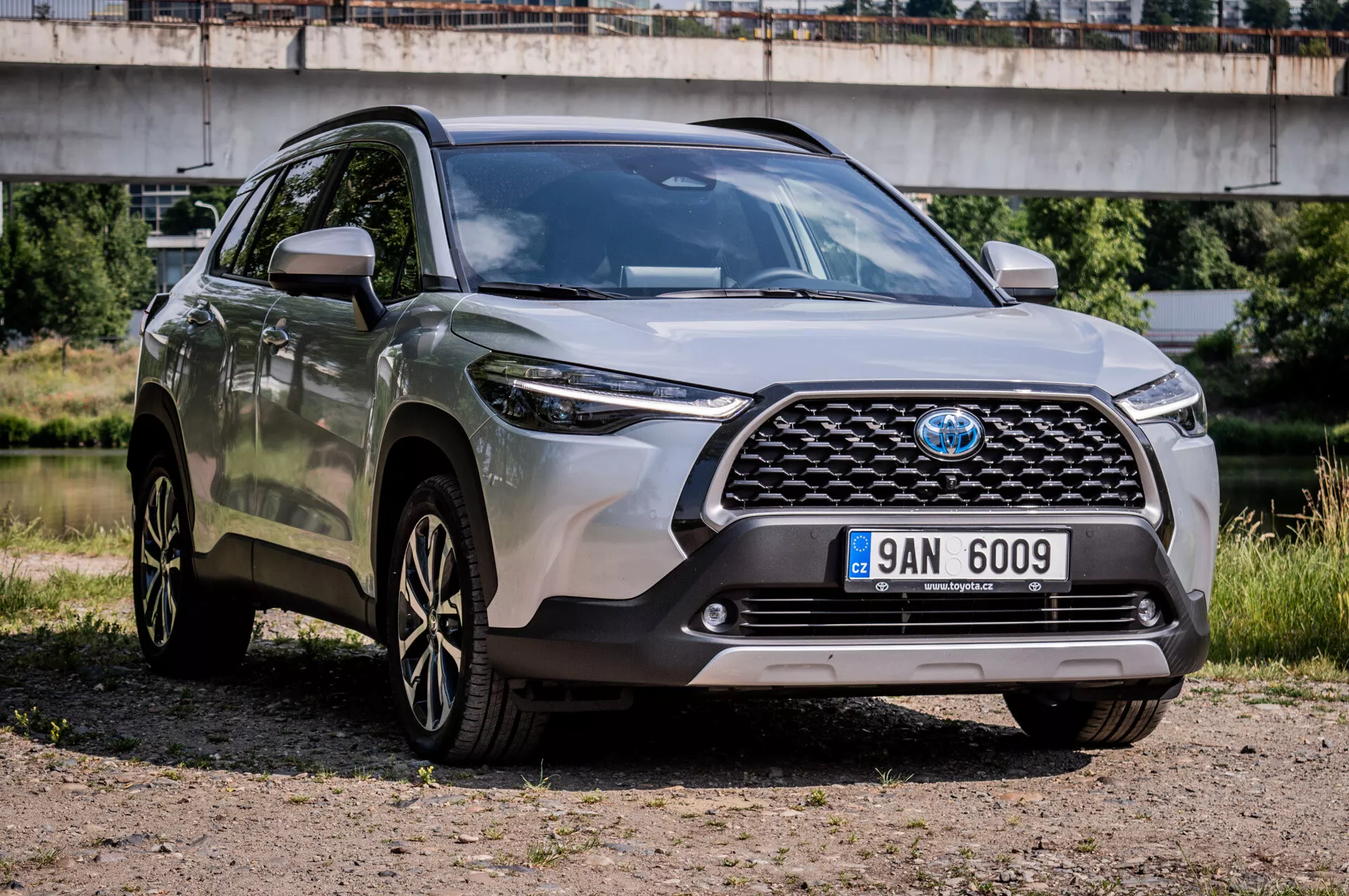 Toyota Corolla Cross 2022 parcheggiata su ghiaia che mostra il suo design robusto, cerchi in lega sportivi a più razze, fari LED avanzati e alta distanza da terra in vista frontale di tre quarti evidenziando la griglia frontale, paraurti e caratteristiche del lato conducente con un ponte sullo sfondo semi-urbano