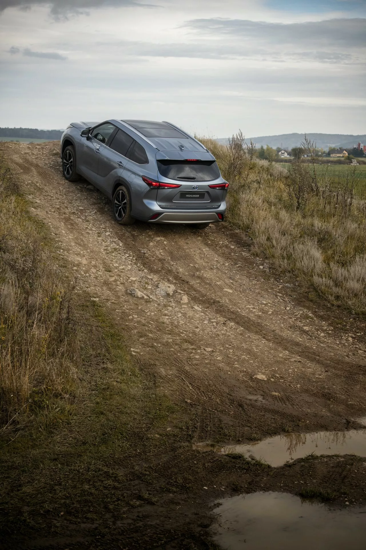 Toyota Highlander scura 2021 catturata da un angolo alto su una strada sterrata, mostrando la vista posteriore da tre quarti, design sportivo e potenziale per viaggi leggeri su tutti i terreni in mezzo a un paesaggio di campagna