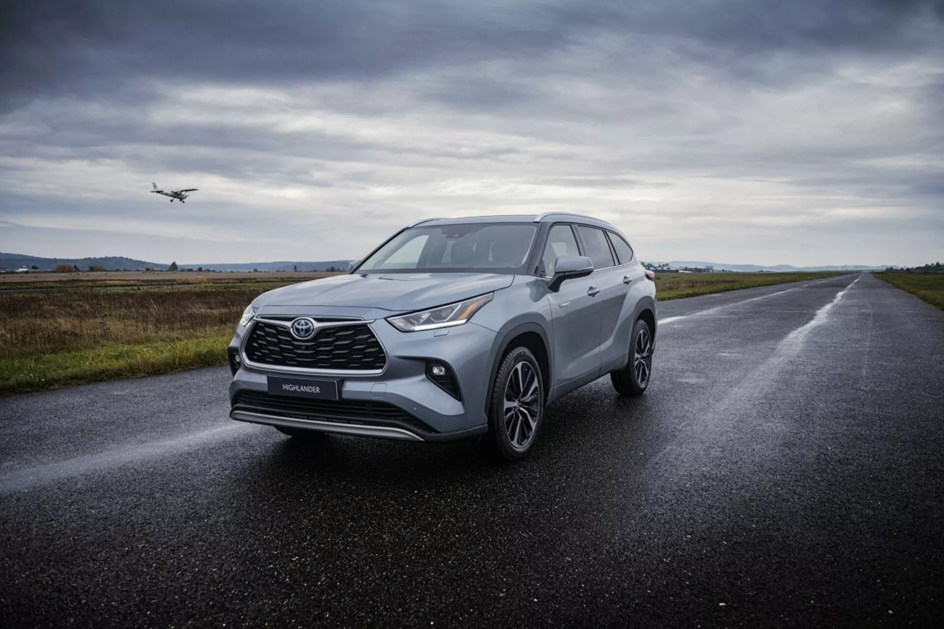 SUV crossover Toyota Highlander 2021 su strada d'asfalto bagnata, mostra i suoi fari angolari, griglia anteriore prominente, ruote in lega a più razze e design aerodinamico sotto una illuminazione diffusa e nuvolosa