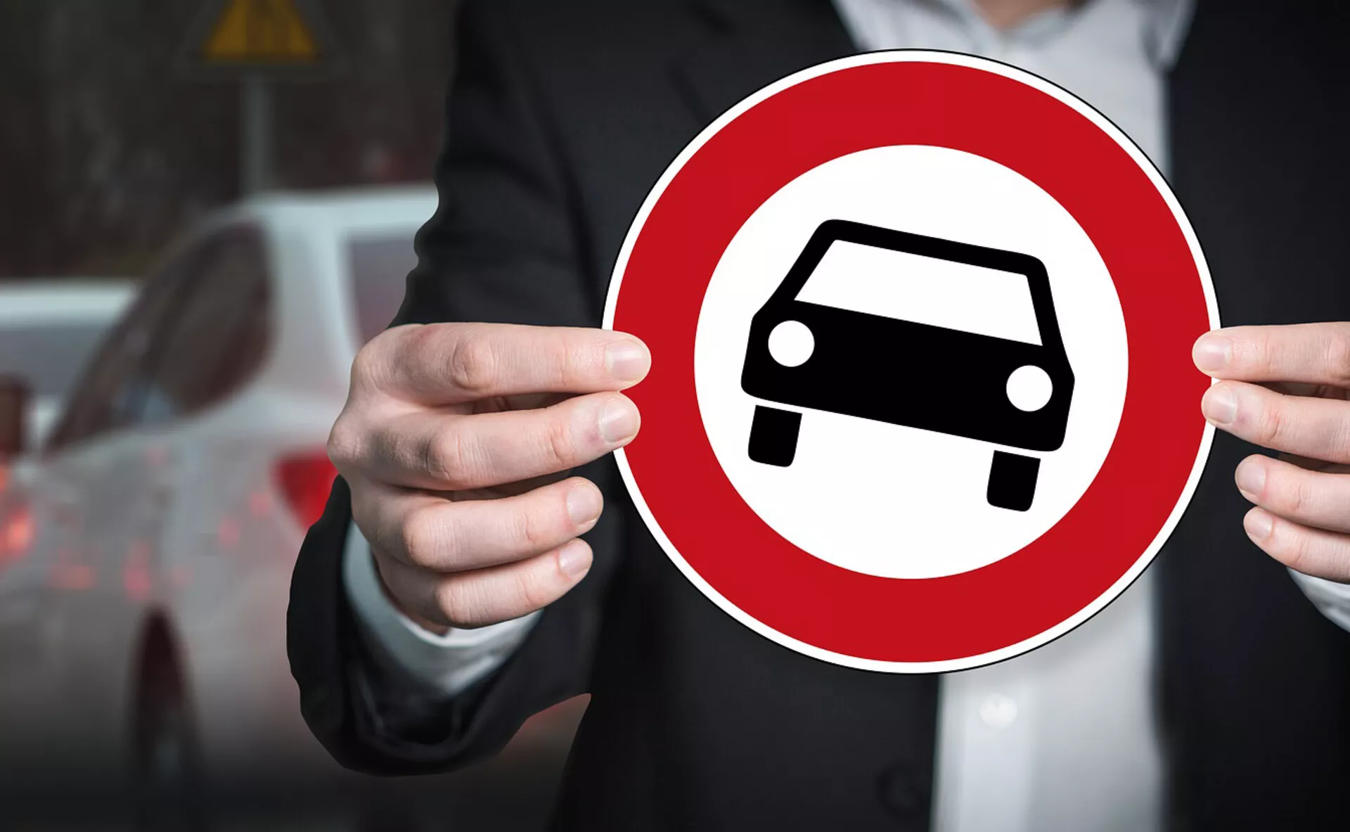 Individuo che tiene un cartello circolare rosso con icona di silhouette di auto bianca, simboleggiando la rappresentazione generica di un'automobile su sfondo urbano sfocato