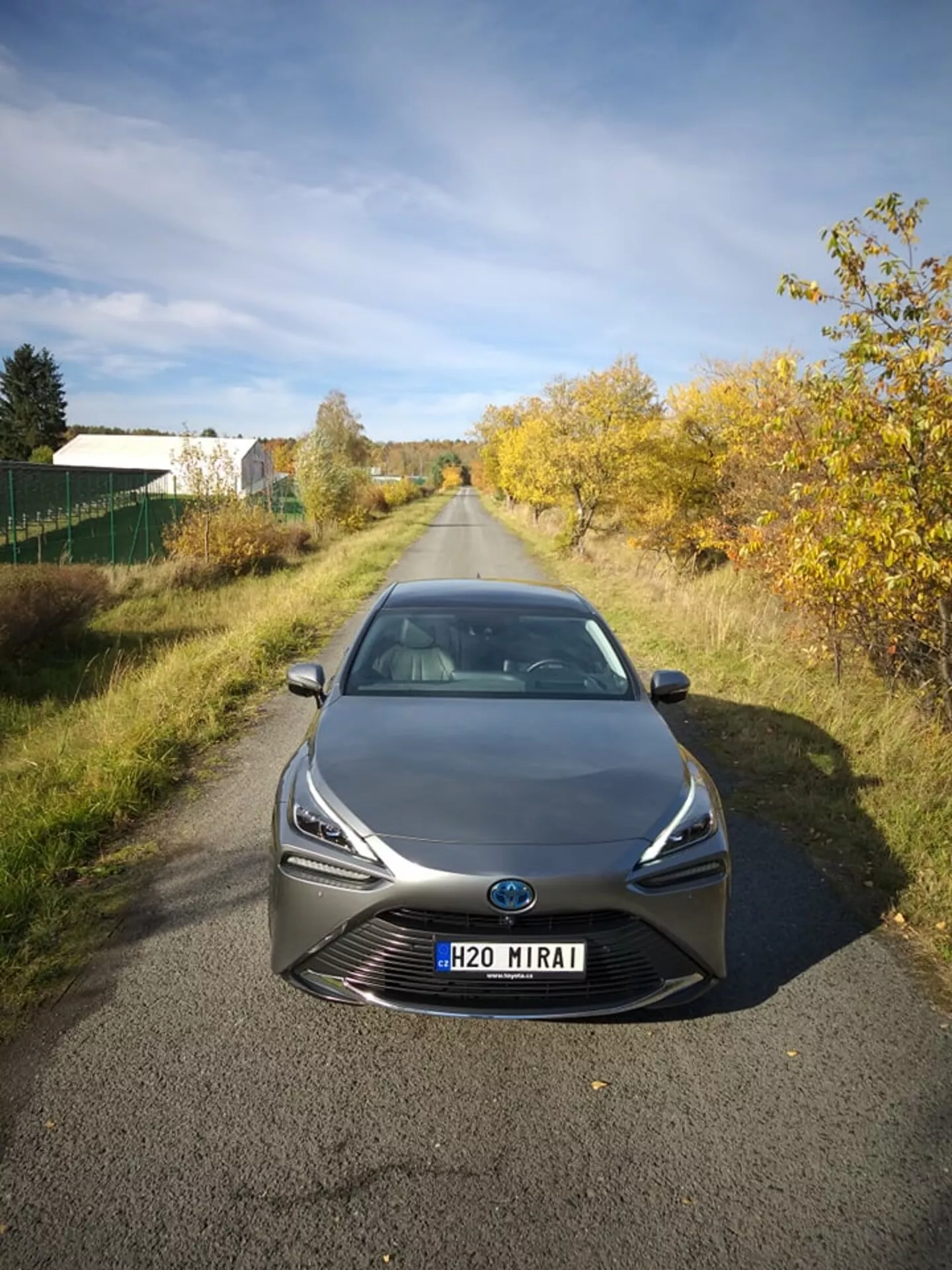 Toyota Mirai 2021 fotografata da un angolo anteriore elevato, mettendo in evidenza i suoi fari LED eleganti e la griglia prominente con emblema centrale che potrebbe includere tecnologia a sensori, ambientata in uno sfondo semi-rurale alla luce naturale, mostrando la tecnologia avanzata delle celle a combustibile a idrogeno e il design aerodinamico