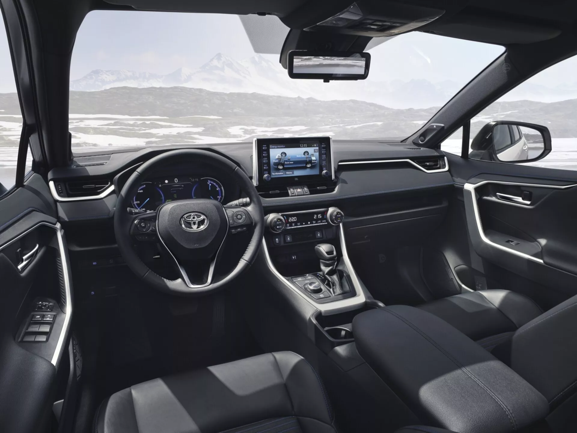 Vista interna del Toyota RAV4 2019 dal sedile posteriore, mostrando il design moderno e le caratteristiche tecnologiche come il volante con pulsanti di controllo, display digitale, sistema di infotainment touchscreen, leva del cambio automatico, sedili tappezzati in scuro, e comandi sul pannello della porta con un paesaggio montano innevato visibile attraverso il parabrezza.