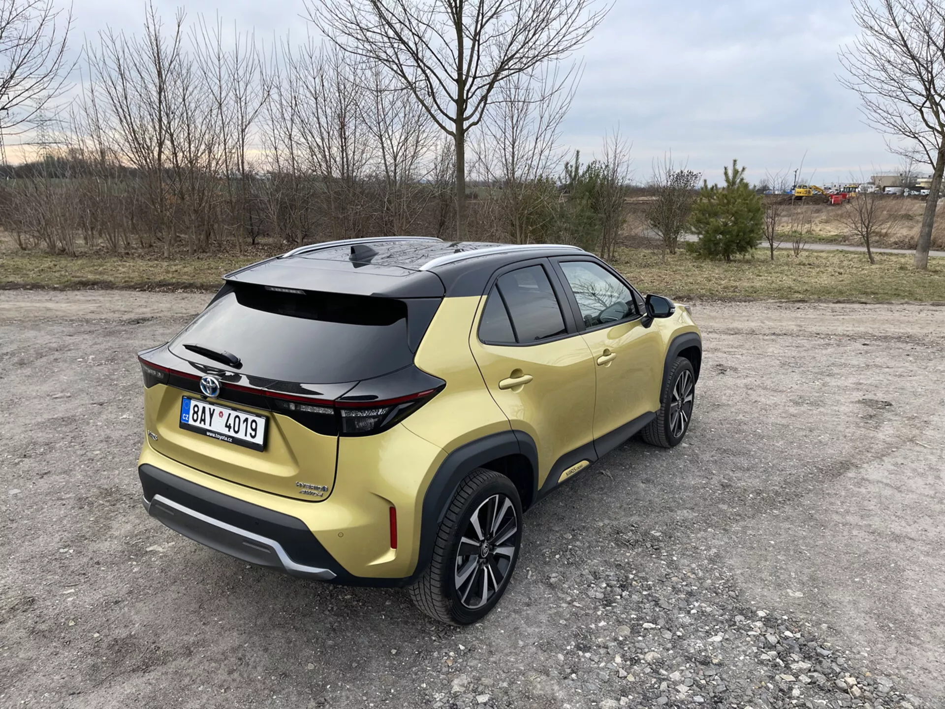 Toyota Yaris Cross 2021 catturata da una vista posteriore di tre quarti che evidenzia il suo spoiler aerodinamico, vernice metallica oro e luci posteriori LED, parcheggiata su una superficie non asfaltata con sfondi naturali ed industriali.