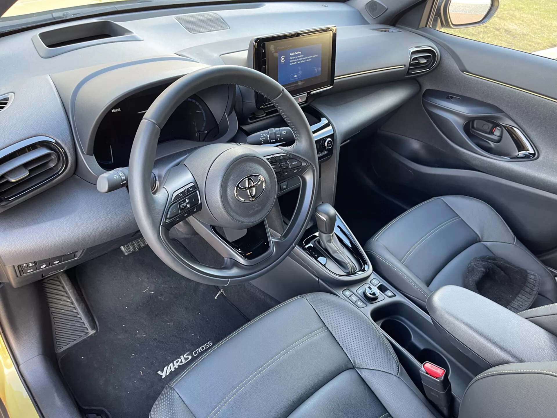 Vista interna della Toyota Yaris-Cross 2021 dai sedili posteriori, evidenziando volante con controlli, bocchette d'aria sul cruscotto, console centrale con sistema di infotainment, controlli del condizionatore, leva del cambio automatico, sedili anteriori a secchiello con aspetto in pelle con branding Yaris Cross, pannelli delle porte con controlli elettrici e design della cabina moderno ed elegante.