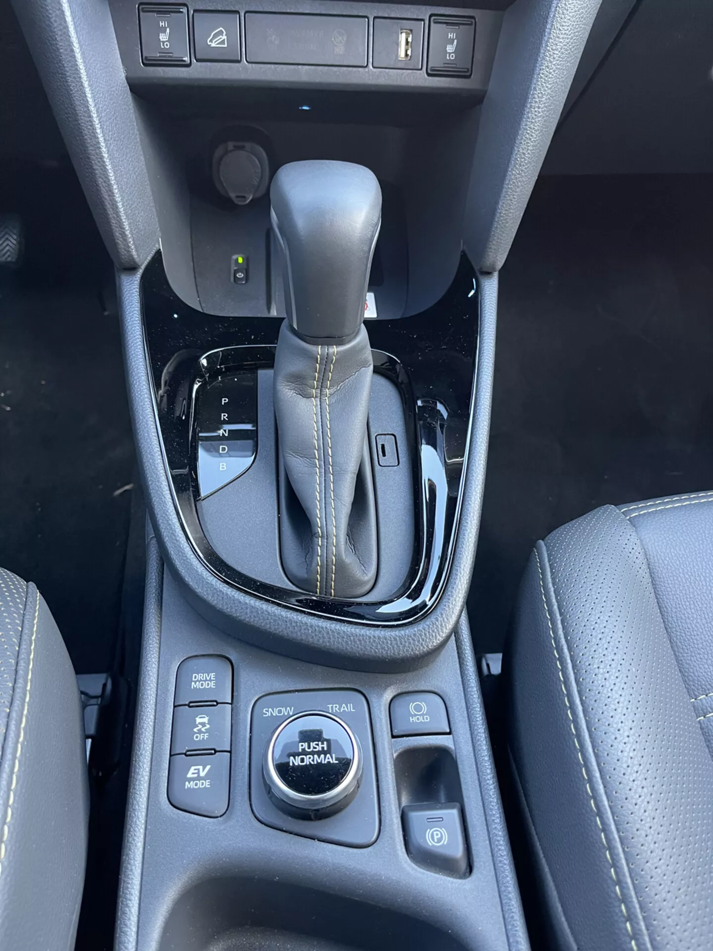 Console centrale interna Toyota Yaris Cross 2021 con leva del cambio automatica in posizione di parcheggio, pulsanti di controllo circostanti per assistenza al parcheggio, mantenimento della frenata, sensori di parcheggio e modalità di guida incluse SPORT, ECO, EV, AUTO e NORMAL, caratterizzata da materiale similpelle con dettagli cuciti e dettagli in nero lucido
