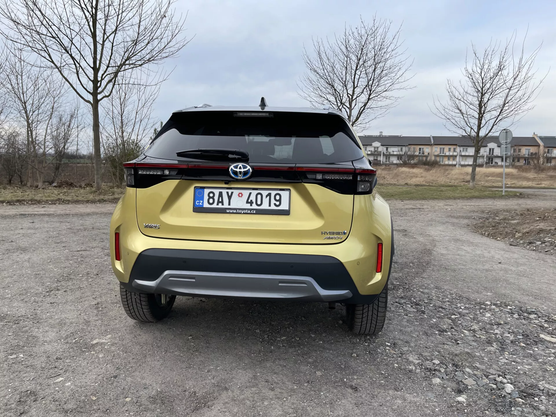 Vista posteriore di una Toyota Yaris Cross gialla 2021 catturata a livello occhi che mette in mostra il suo design moderno, assemblaggio luci posteriori orizzontali, emblema Toyota, nome del modello Urban Cruiser, targa, paraurti posteriore dal design a contrasto con sensori di parcheggio integrati e maniglia del portellone su uno sfondo suburbano.