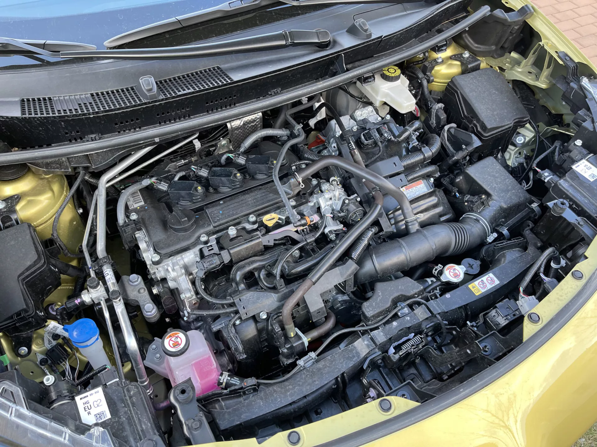 Vista dettagliata del vano motore di una Toyota Yaris-Cross 2021 con il cofano aperto che mostra il blocco motore a quattro cilindri, sistema di aspirazione dell'aria, batteria del veicolo, serbatoio del refrigerante e vari contenitori di fluidi