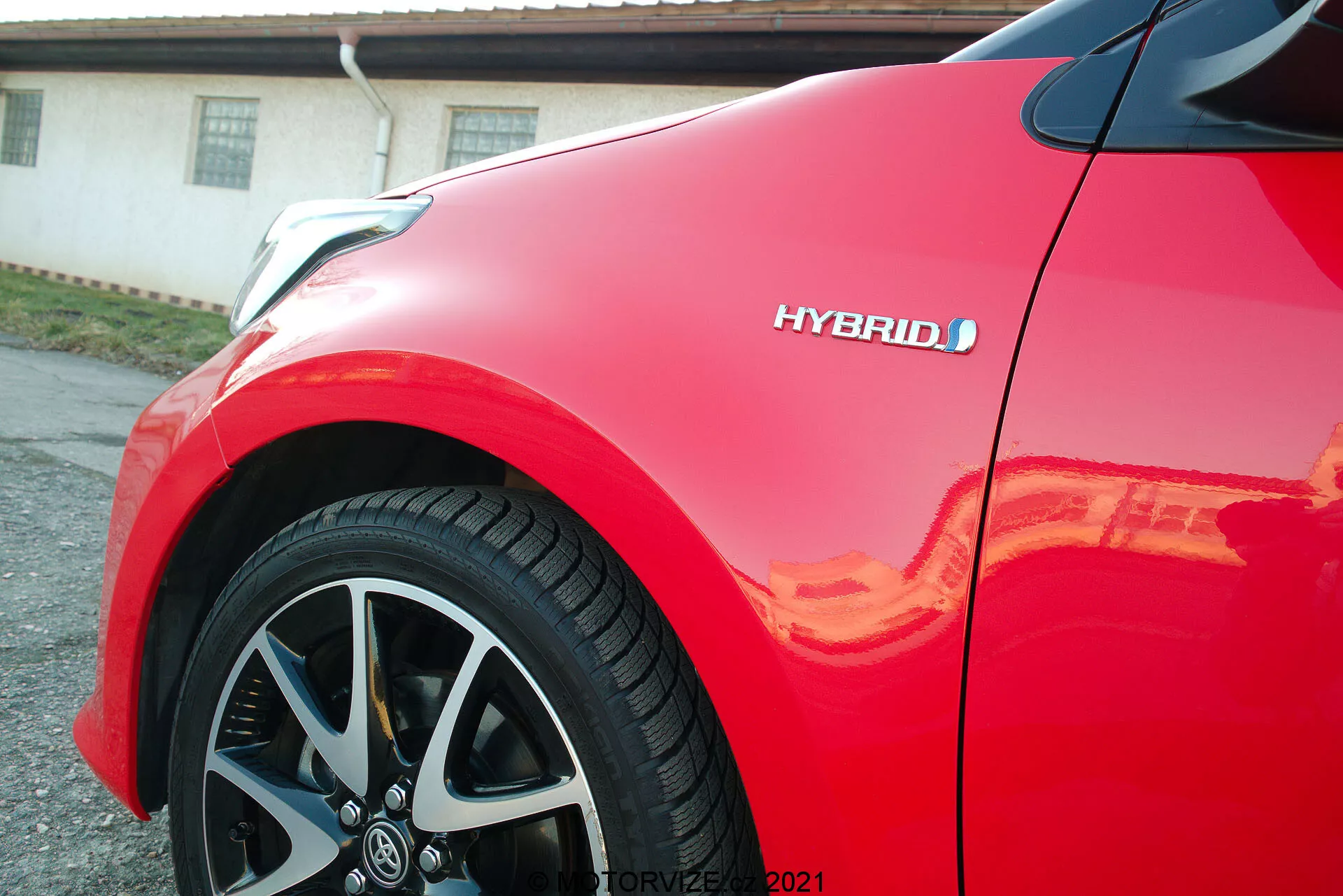 Vista ravvicinata di una Toyota Yaris 2020 rossa, con attenzione al pannello anteriore e alla ruota in lega a più razze. Il badge ibrido è visibile, evidenziando la tecnologia ecologica del veicolo. La vernice lucida riflette la luce del sole, enfatizzando la finitura di alta qualità e il design aerodinamico e moderno dell'auto.