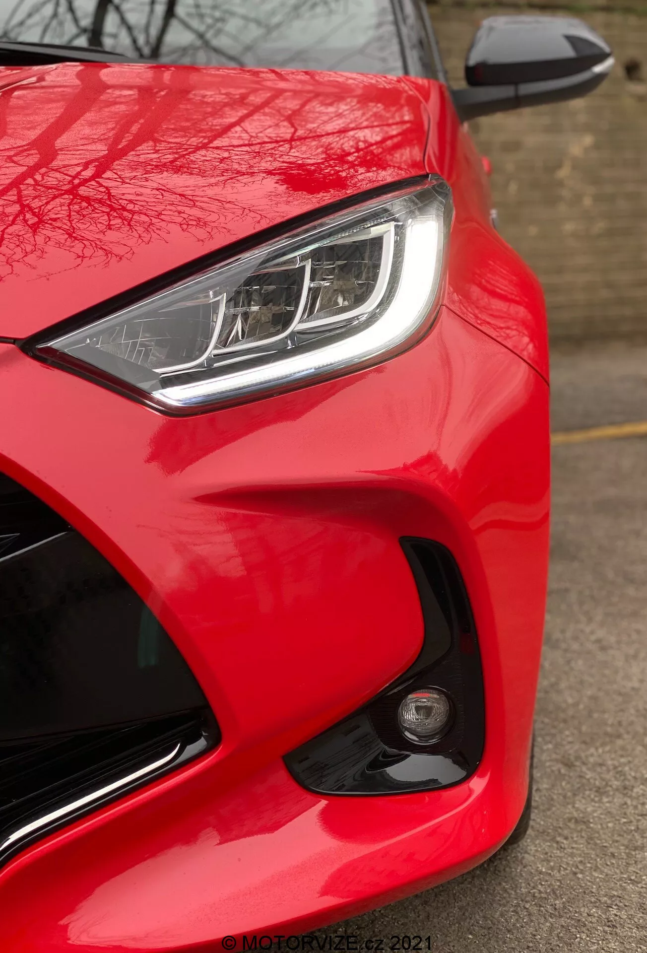 Vista ravvicinata della Toyota Yaris 2020, inquadrata dal lato anteriore sinistro da un'angolazione bassa. L'immagine mette in evidenza il colore rosso brillante e lucido dell'auto, il faro LED o HID elegante con lampade diurne distintive e lo specchietto laterale dello stesso colore della carrozzeria parzialmente visibile con indicatore di direzione integrato. Il design aerodinamico è enfatizzato dalle linee scolpite e dalle curve attorno al faro e al cofano. Un faro antinebbia rotondo o un sensore sul paraurti anteriore suggeriscono caratteristiche di sicurezza avanzate. La mancanza di sfocatura indica che l'auto è ferma e una filigrana 'MOTORZETZ. 2021' è visibile in basso a destra.