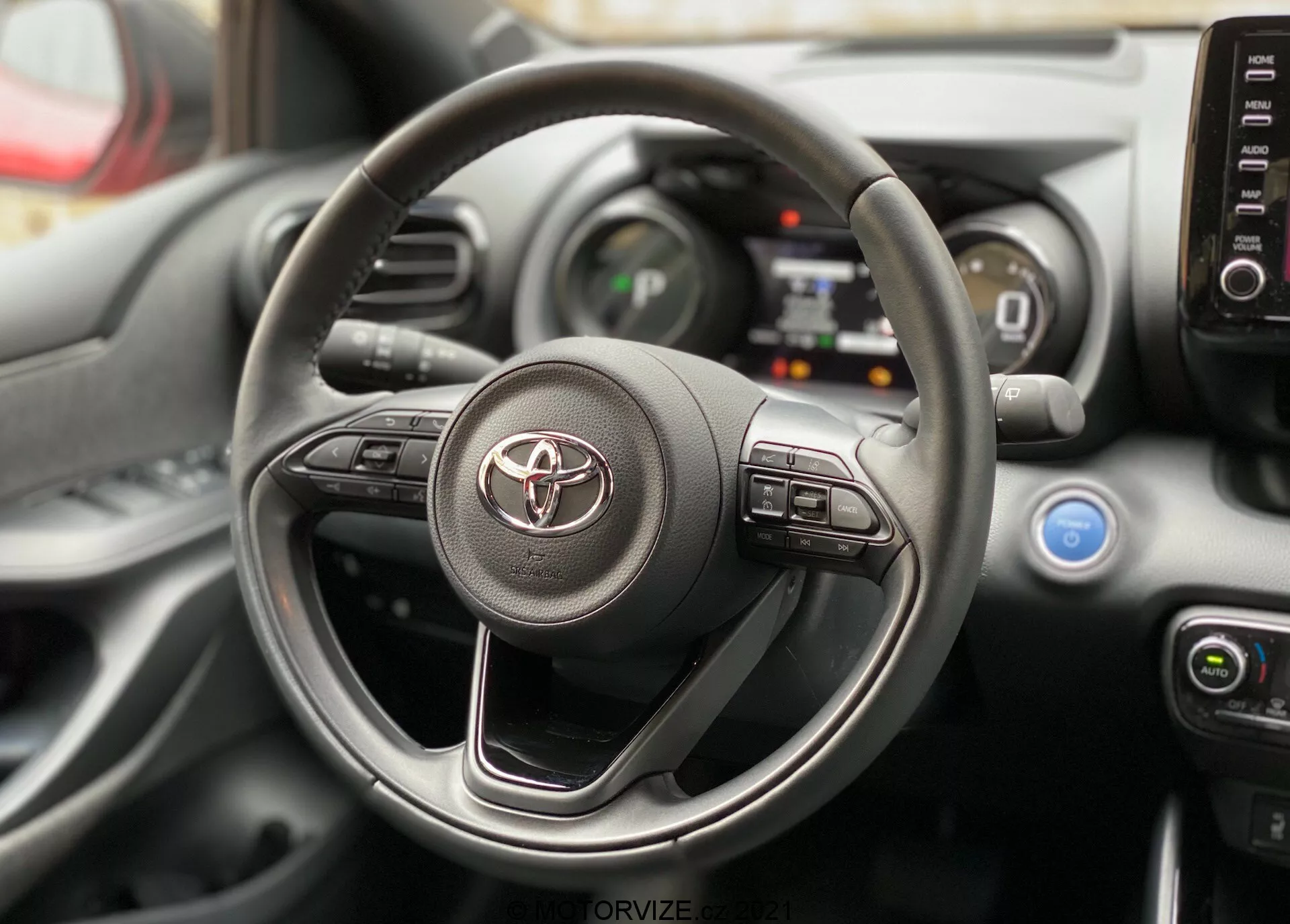 Vista interna di una Toyota Yaris 2020 con focus sul volante e il cruscotto dalla prospettiva del posto di guida. Il volante presenta il logo Toyota e controlli visibili per il sistema di infotainment e i sistemi di assistenza alla guida. Dietro di esso, sono visibili parti del quadro strumenti digitale e della console centrale con manopole di controllo del clima e bocchette di aerazione.