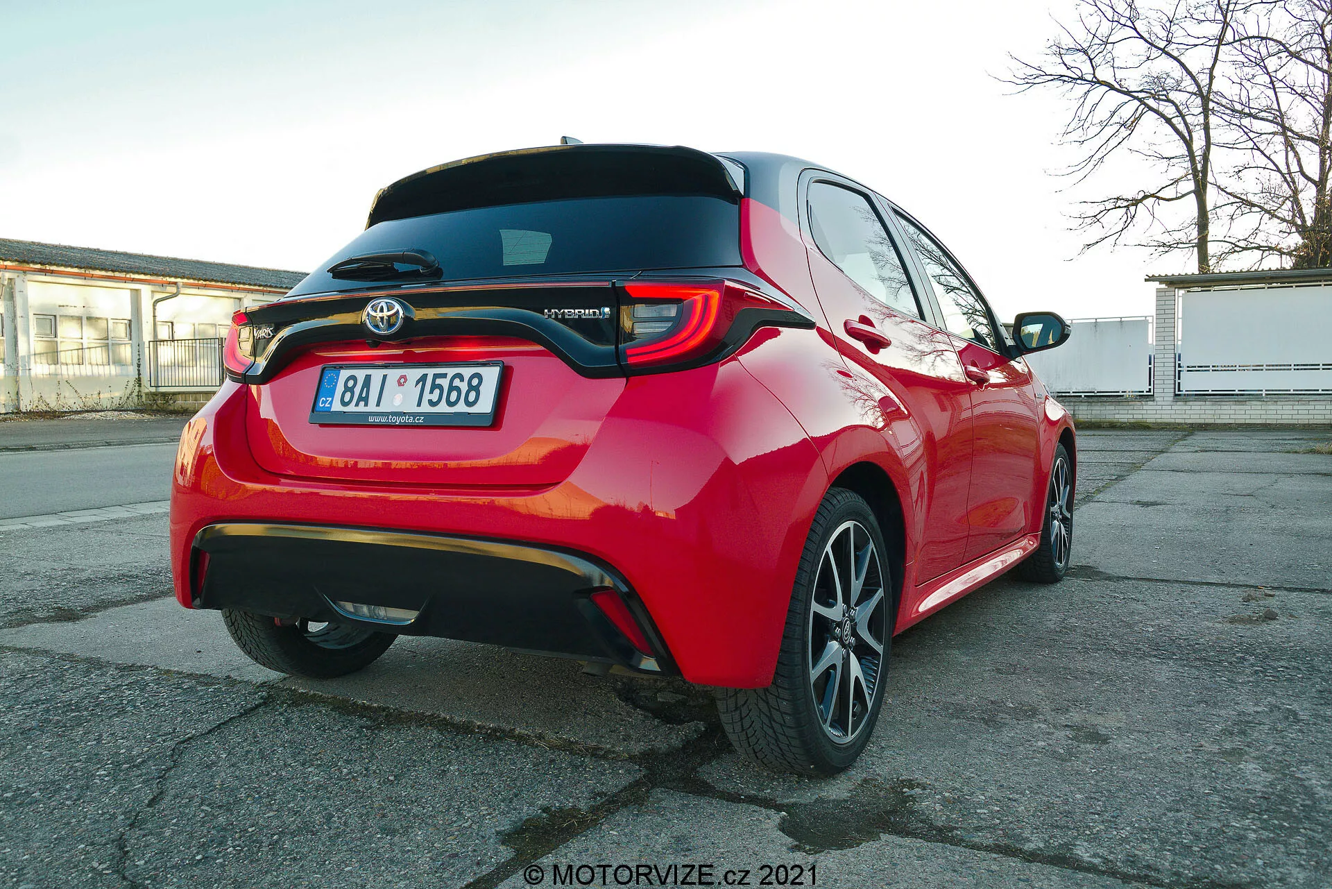 Vivace auto passeggeri Toyota Yaris 2020 rossa fotografata da un'angolazione posteriore a tre quarti, che enfatizza le luci posteriori a LED, lo spoiler posteriore, il distintivo del modello ibrido, cerchi in lega a più razze e funzioni visibili di assistenza alla guida come i sensori di parcheggio e la telecamera posteriore, catturata da una prospettiva leggermente inferiore all'altezza dell'auto.