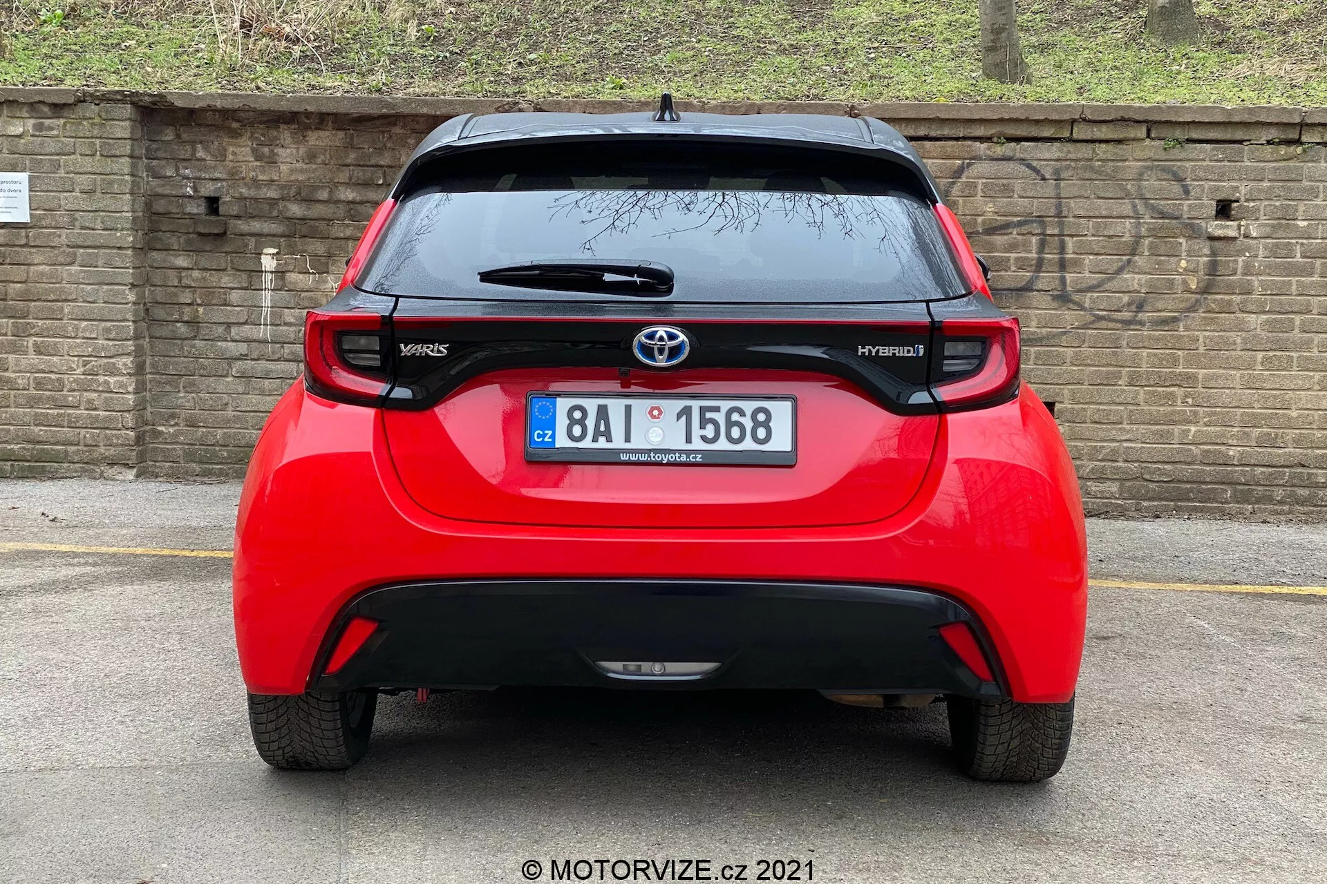 Vista posteriore di una Toyota Yaris Hybrid rossa del 2020, centrata e scattata a un angolo diretto. L'immagine rivela l'intera larghezza del veicolo, mostrando l'emblema Toyota, i nomi dei modelli 'Yaris' e 'Hybrid', uno spoiler, un'antenna montata sul tetto, una striscia di luce del freno in alto e luci posteriori distintive. Sono inoltre visibili il lunotto con un tergicristallo e una parte inferiore del paraurti che potrebbe ospitare sensori o riflettori. Lo sfondo include un muro di mattoni o una recinzione e della vegetazione, suggerendo un'auto parcheggiata più in basso o un angolo della fotocamera più basso.