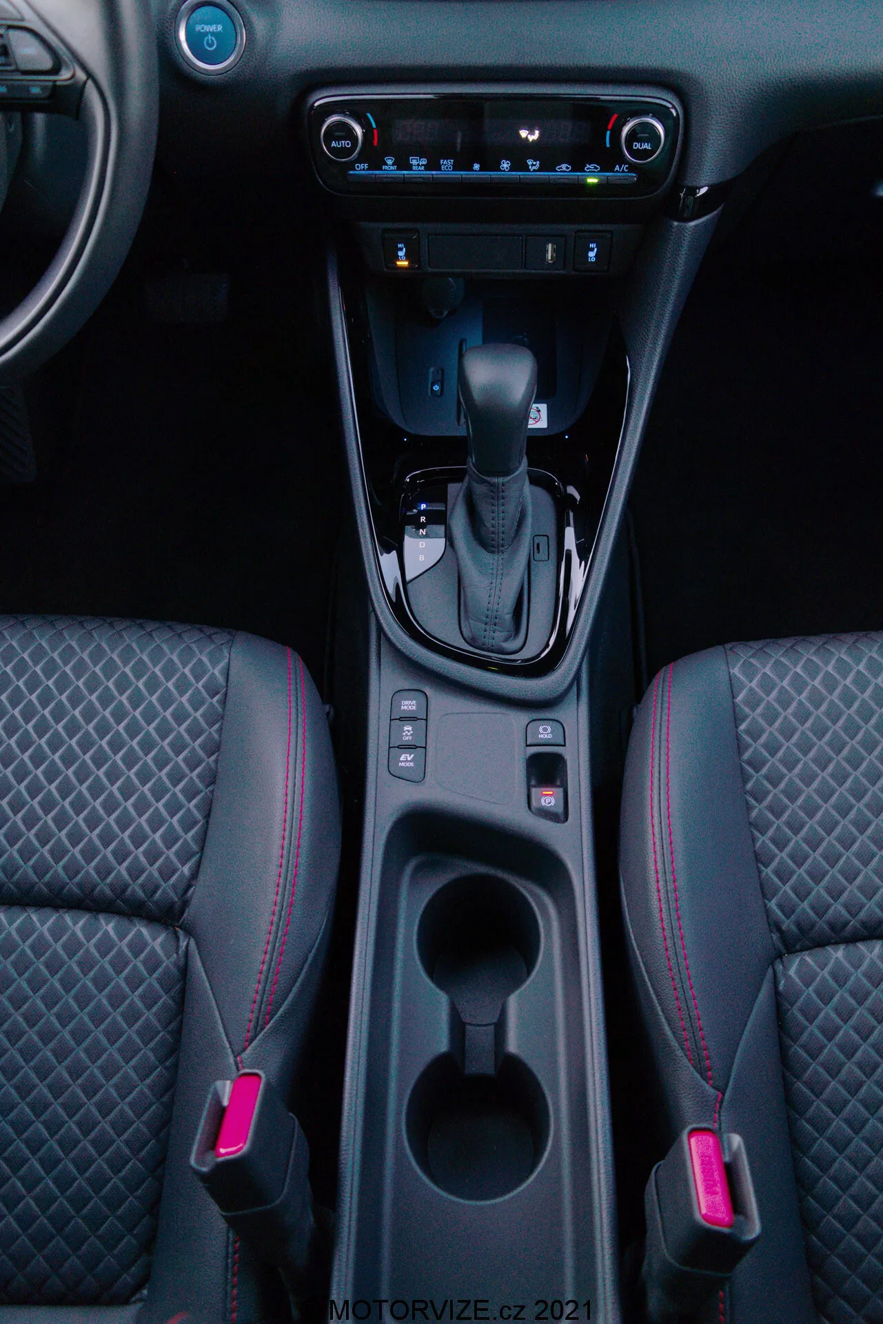 Ripresa verticale dell'interno di una Toyota Yaris 2020 con attenzione alla console centrale, con leva del cambio in posizione 'P', pulsante del freno di stazionamento elettronico, due portabicchieri illuminati, spazio di stivaggio, controllo climatizzazione digitale, sistema multimediale touchscreen, porta USB, bocchette d'aria e sedili anteriori bicolore con cuciture a motivo diamante e cuciture accentate rosse.