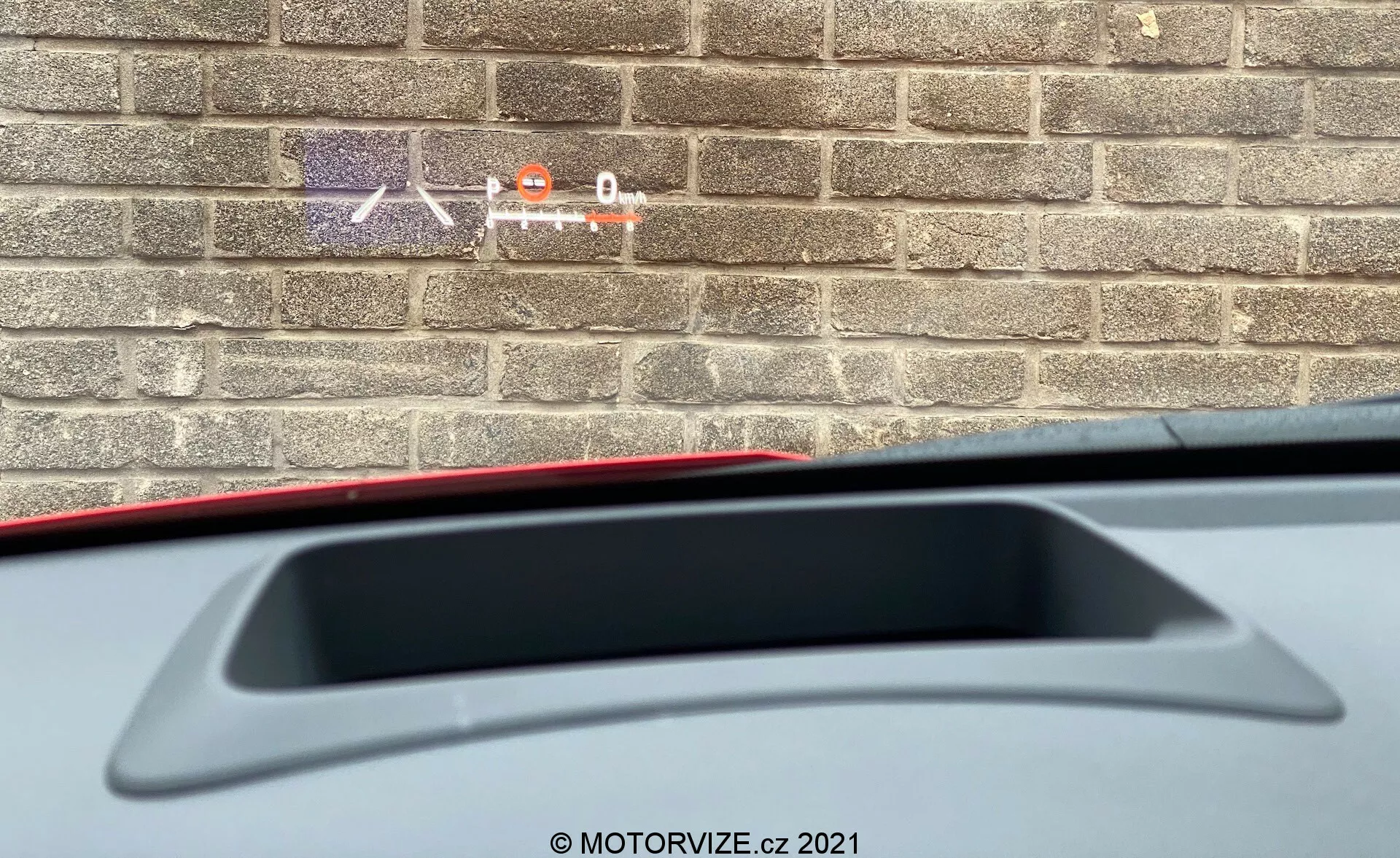 Vista ravvicinata dall'interno di una Toyota Yaris 2020 che mostra una prospettiva parziale dal sedile del conducente verso la parte posteriore. L'attenzione è sulla mensola posteriore con una luce di stop alta e uno spoiler posteriore integrato. L'immagine mostra anche parte del materiale degli interni mentre un muro di mattoni all'esterno suggerisce che l'auto sia parcheggiata vicino a un edificio.