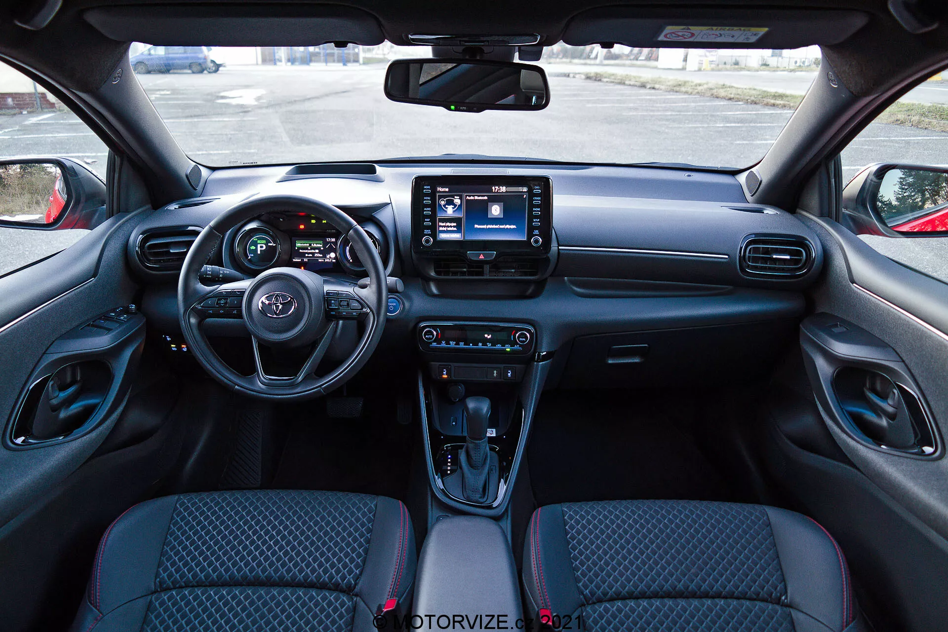 Vista dell'interno di una Toyota Yaris 2020 dalla prospettiva del sedile del conducente, con un cruscotto moderno che presenta un volante con comandi integrati, un quadro strumenti parzialmente visibile con schermi digitali e un sistema di infotainment centrale con interfaccia touchscreen. Il cruscotto comprende le bocchette dell'aria e le manopole di controllo del clima, mentre la console centrale presenta comandi per le modalità di guida, una leva del cambio automatico con copertura simile alla pelle, portabicchieri e un vano portaoggetti. I sedili con rivestimento in tessuto hanno un design a motivi e i pannelli interni delle porte includono comandi per i finestrini elettrici e la regolazione degli specchietti.