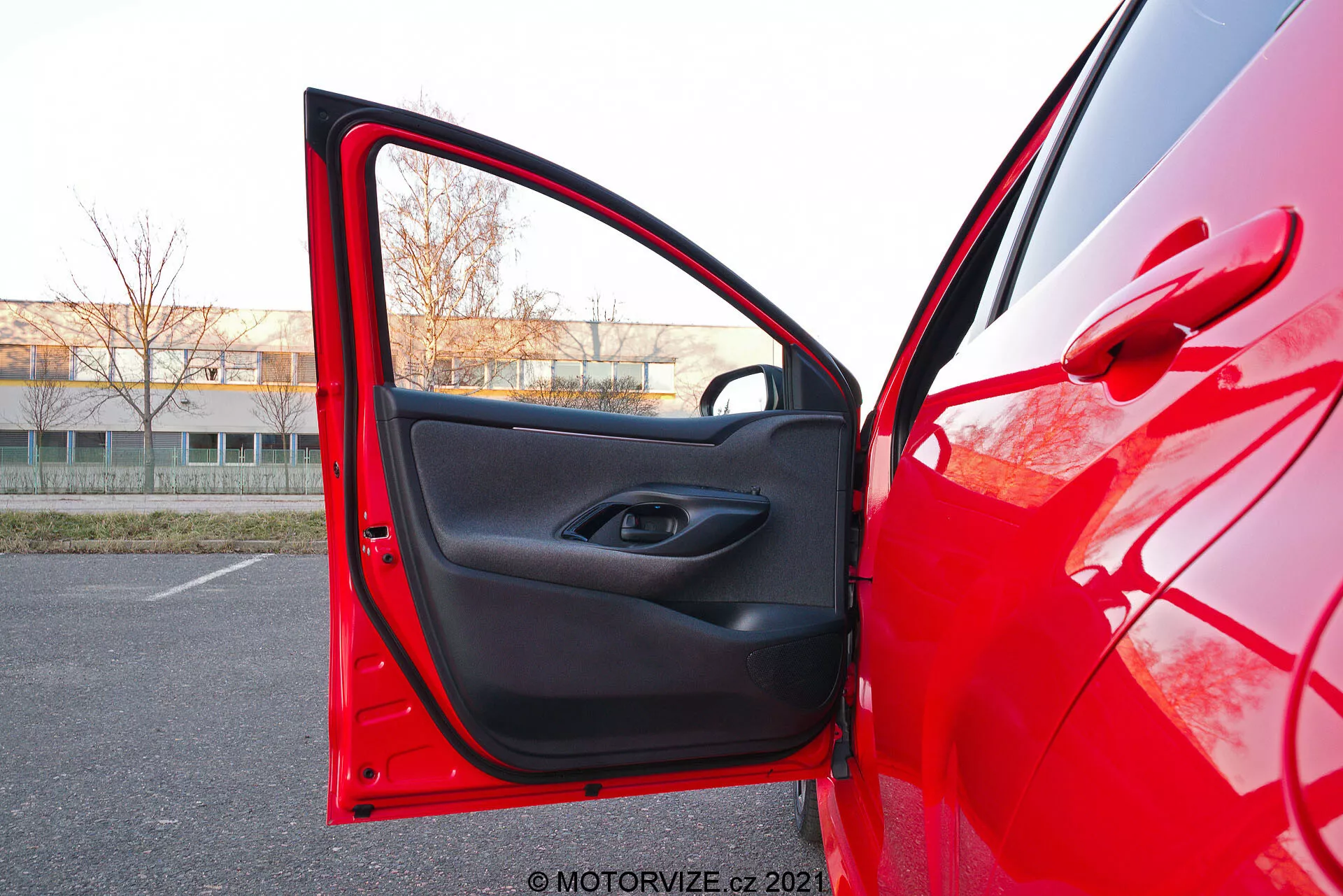 Vista ravvicinata del lato conducente di una Toyota Yaris 2020 rossa con la portiera anteriore aperta, catturata da un angolo leggermente basso. La telecamera è posizionata all'esterno, vicino alla porta, puntando verso l'interno, mostrando la vernice esterna rossa brillante che riflette la luce e il rivestimento interno nero con un design minimalista. La porta ha un pulsante di controllo del finestrino elettrico all'interno dell'incavo della maniglia e una tasca portaoggetti sotto.