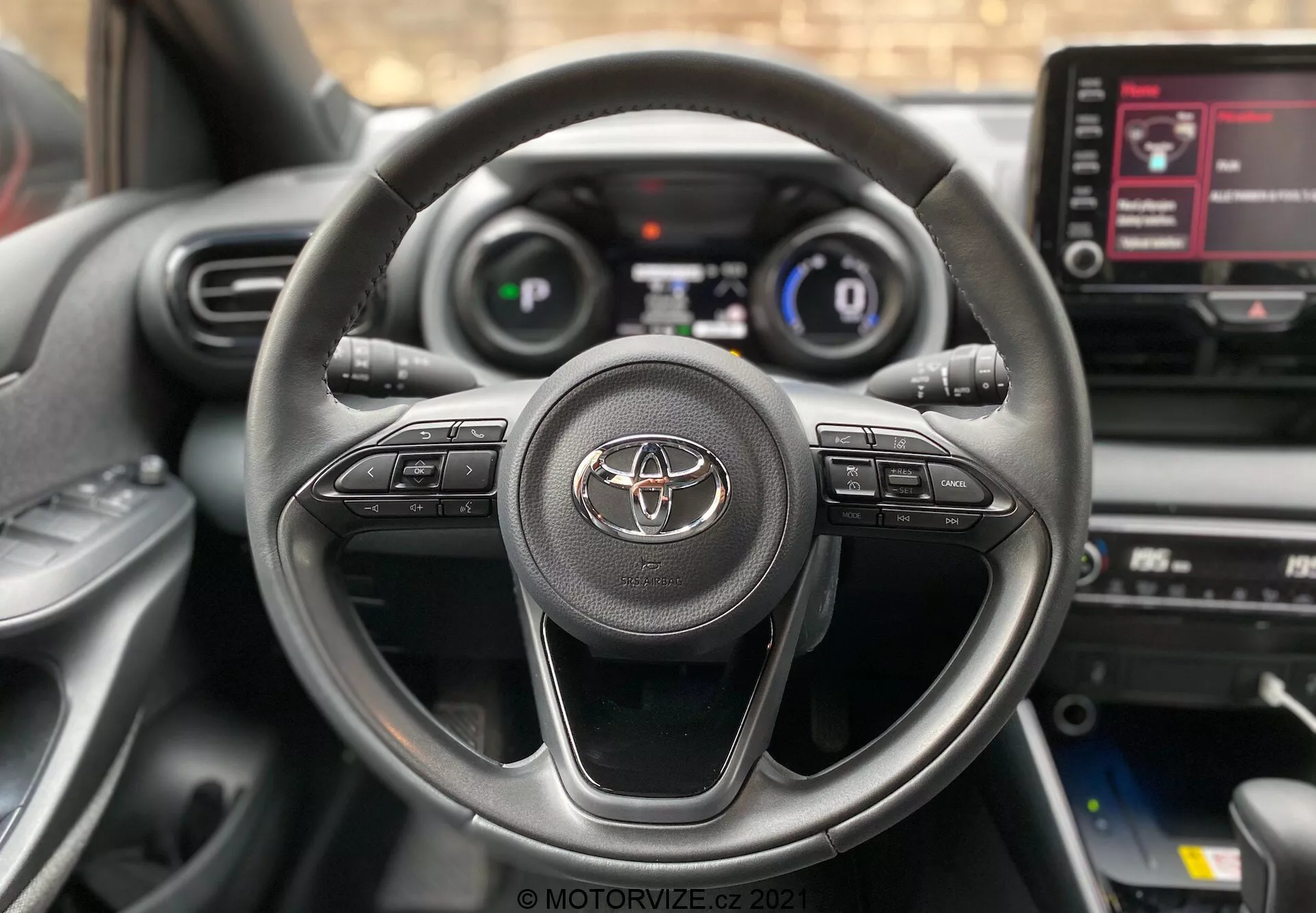 Primo piano dell'interno della Toyota Yaris 2020 dal sedile del conducente, con volante a tre razze con emblema Toyota e pulsanti di controllo per audio, chiamate hands-free e controllo della velocità. Parzialmente visibile il quadro strumenti digitale che mostra la velocità e le informazioni essenziali di guida. È visibile anche la console centrale con touchscreen di infotainment che mostra le opzioni del menu.