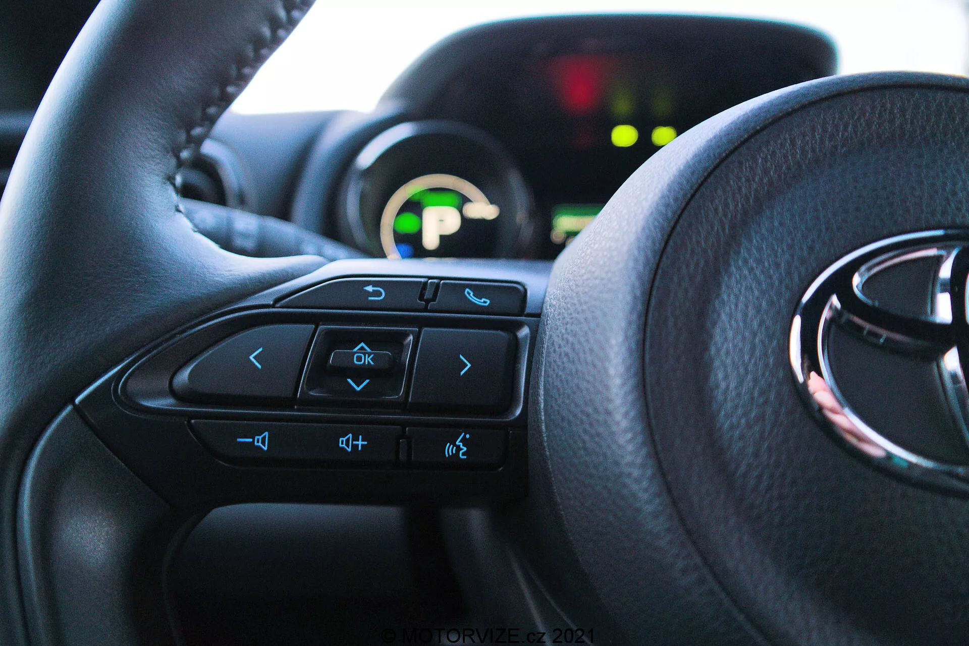Vista interna ravvicinata di una Toyota Yaris 2020 dalla prospettiva del sedile del conducente, con particolare attenzione al volante con comandi multifunzione e al cruscotto sullo sfondo. Il volante domina i due terzi inferiori dell'immagine, con frecce, comando vocale, volume e pulsanti del sistema di intrattenimento visibili. Il cruscotto parzialmente visibile è illuminato con un display digitale verde ed è fuori fuoco a causa della bassa profondità di campo. La luce naturale illumina l'interno, enfatizzando il comfort e un'interfaccia user-friendly.