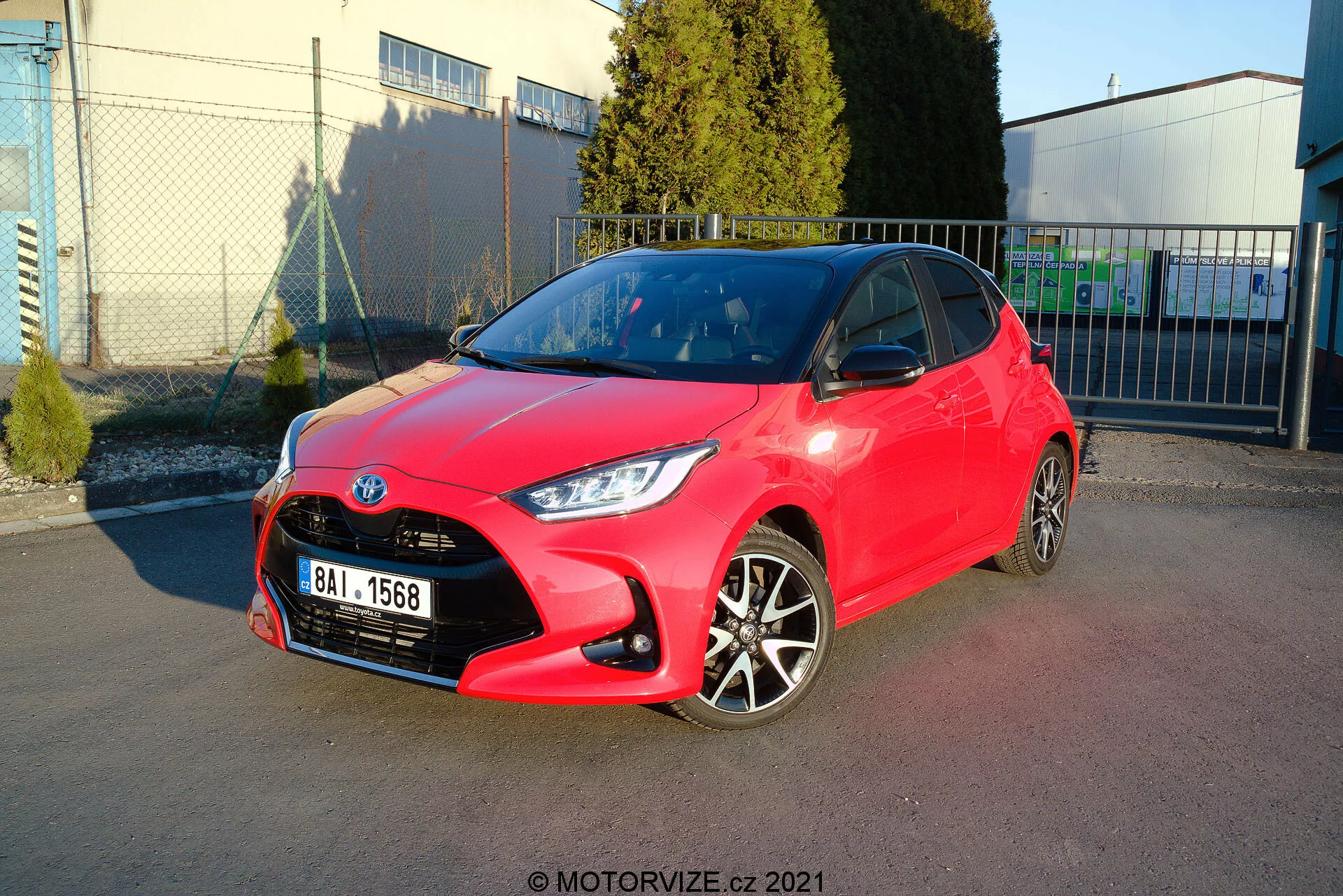 Immagine di una Toyota Yaris hatchback del 2020 di colore rosso brillante parcheggiata in diagonale, mostrando sia il profilo anteriore che quello laterale. Il design dei fari è affilato e angolare, la griglia ha un motivo a nido d'ape con un emblema centrale e il paraurti anteriore presenta contorni visibili con luci fendinebbia. Il profilo laterale rivela cerchi in lega bicolore, maniglie delle porte e specchietti in tinta con la carrozzeria, con linee e contorni netti lungo il lato. La vernice lucida suggerisce una condizione nuova o ben mantenuta. Lo sfondo include un cielo limpido, strutture industriali e ombre morbide, indicando un ambiente all'aperto, probabilmente un'area di parcheggio vicino a una struttura commerciale o industriale.