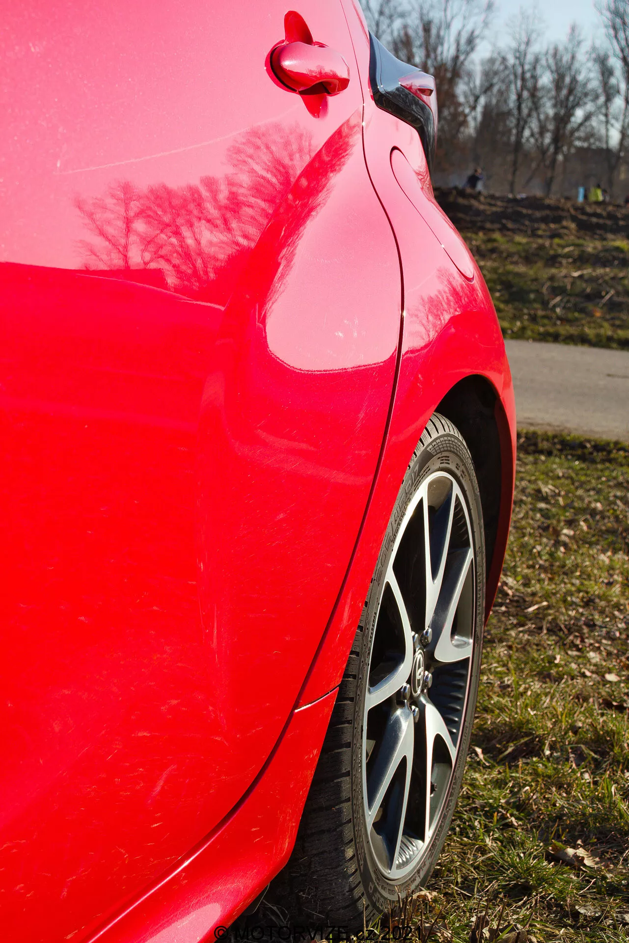Vista laterale ravvicinata del pannello posteriore del Toyota Yaris 2020 rosso scattata da un'angolazione leggermente verso l'alto che evidenzia la finitura rossa brillante, il cerchione in lega multirazze, il battistrada del pneumatico, la curvatura pulita del pannello posteriore e del paraurti, il riflesso lucido della vernice con alberi e un cielo azzurro chiaro, e il tappo del serbatoio e il dettaglio della rifinitura visibili.