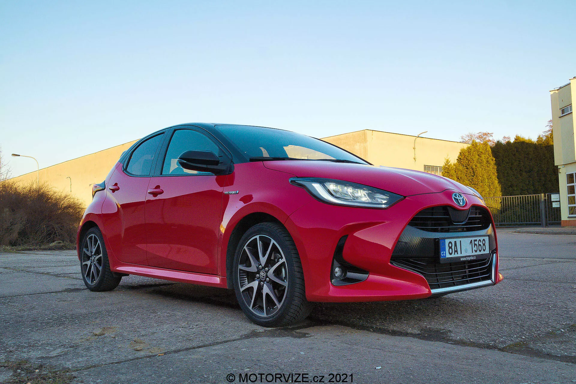 Vista frontale a tre quarti dal basso di una vivace Toyota Yaris hatchback 2020 rossa che mostra fari a LED o HID angolari e nitidi, griglia prominente con emblema centrale, cerchi in lega a più razze, indicatori di direzione integrati negli specchietti laterali, superfici riflettenti pulite e un possibile sistema frenante orientato alle prestazioni. Scattato in un'area di parcheggio industriale illuminata caldamente con il watermark motorvize.cz 2021.