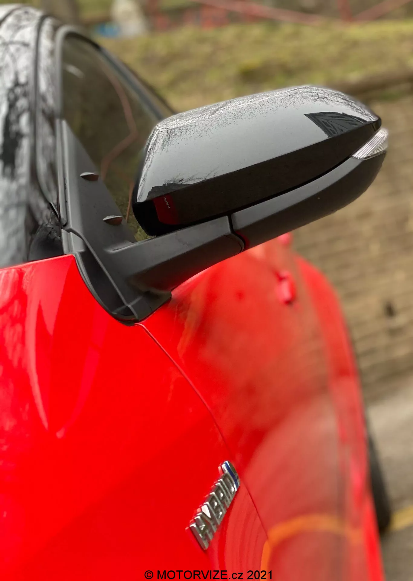 Primo piano dello specchietto laterale dispiegato di una Toyota Yaris 2020, con un design aerodinamico nero che contrasta con la carrozzeria rossa lucida della vettura. Lo specchio mette in risalto possibili caratteristiche tecnologiche come la regolazione elettrica, il riscaldamento e gli indicatori di svolta integrati, anche se le caratteristiche specifiche non sono confermate. Le gocce d'acqua suggeriscono pioggia recente o lavaggio dell'auto, mentre uno sfondo sfocato e una profondità di campo ridotta mantengono l'attenzione sullo specchio. Una parte dell'emblema sul parafango anteriore è visibile ma sfocata.
