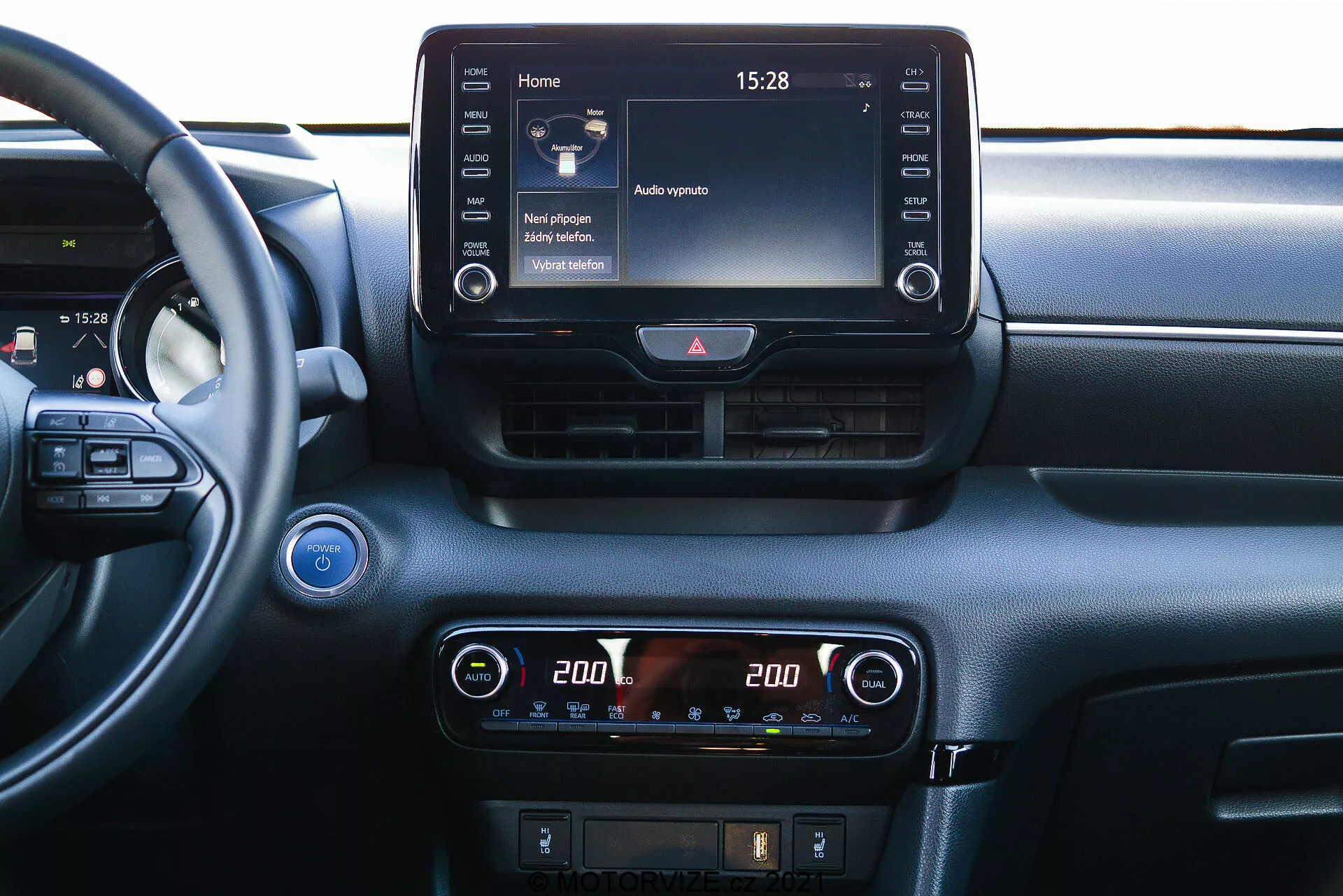 Vista ravvicinata del cruscotto centrale di una Toyota Yaris 2020 da una prospettiva interna. Il volante è parzialmente visibile a sinistra, suggerendo un angolo di visione dal sedile passeggero o centrale. Il cruscotto presenta un touchscreen di infotainment prominente in cima alla console centrale, che mostra una schermata principale con diverse icone e l'orario '15:28'. Sotto di esso, un display di controllo del clima digitale con impostazioni di temperatura a doppia zona, ciascun lato impostato su '20.0' gradi Celsius. I controlli aggiuntivi per lo sbrinamento e potenzialmente l'assistenza alla guida sono visibili sotto i controlli del clima. Sopra lo schermo di infotainment, ci sono due prese d'aria orizzontali con un pulsante delle luci di emergenza in mezzo. Un pulsante di avvio/arresto è parzialmente visibile sull'estremità sinistra, indicando una tecnologia di accensione senza chiave. Una griglia dell'altoparlante si trova nell'angolo in alto a sinistra. Il materiale del cruscotto presenta elementi sia lisci che testurizzati, suggerendo un focus sul comfort e la tecnologia.