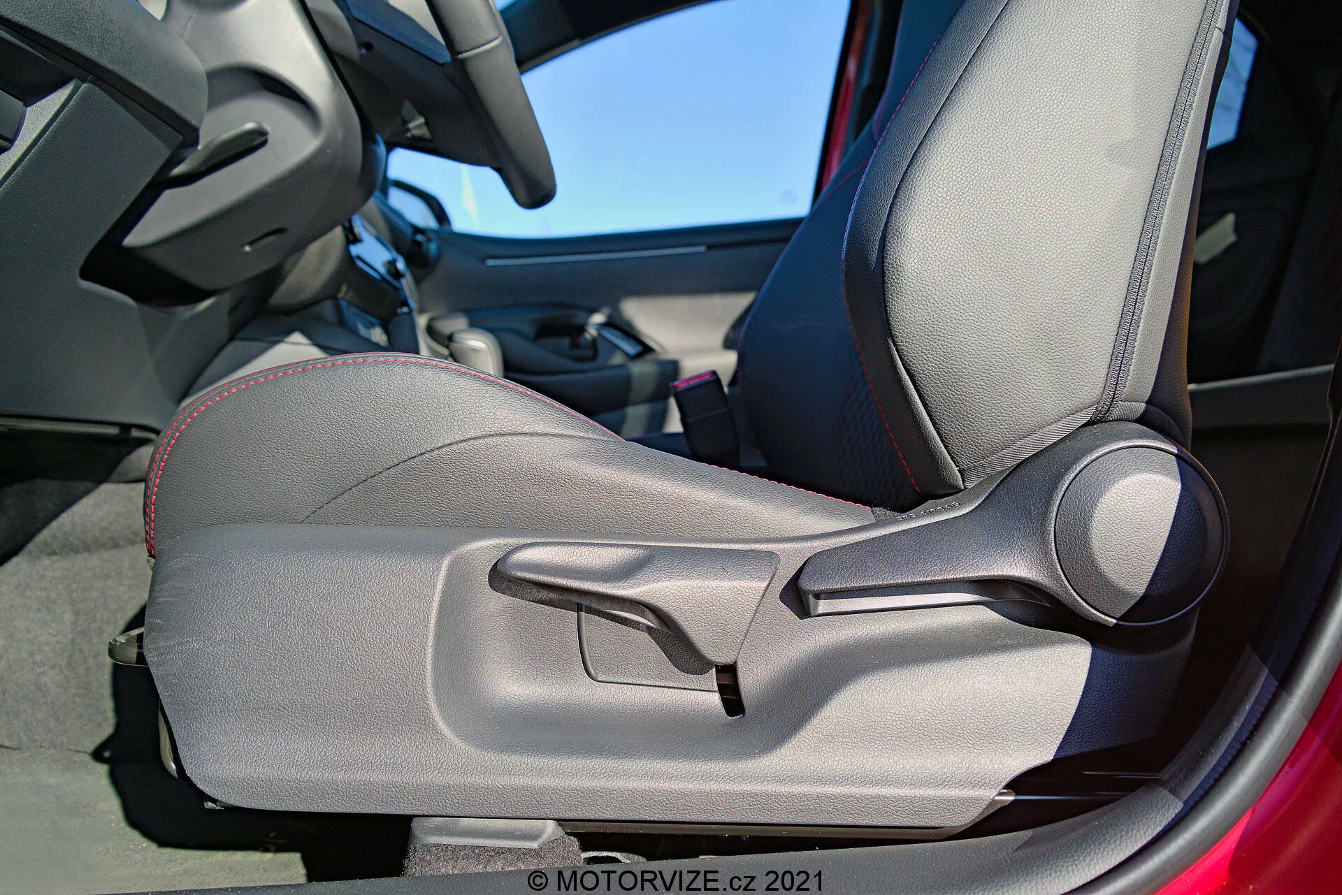 Scatto dettagliato interno della Toyota Yaris 2020 focalizzato sulla porta e sul sedile del conducente, evidenziando il rivestimento bicolore, le cuciture decorative, il design sagomato per il supporto ergonomico, i fianchetti regolabili manualmente e una prominente leva di regolazione manuale del sedile. Il telaio della porta è parzialmente visibile, suggerendo che la porta sia aperta.