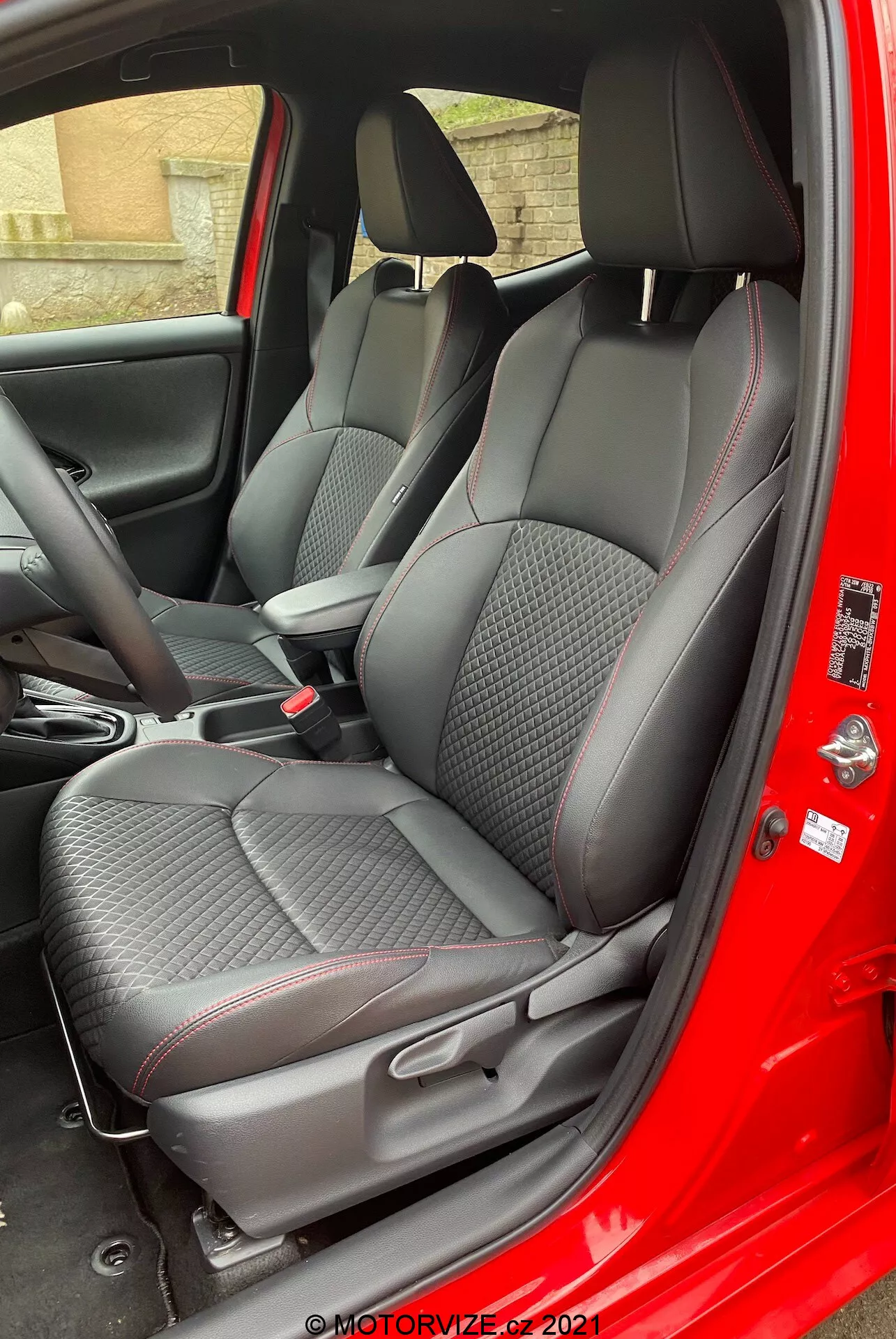 Interno della Toyota Yaris 2020 visto dal lato del passeggero con la porta aperta, evidenziando sedili in tessuto premium con motivi a diamante e dettagli rossi, poggiatesta regolabili, comandi della console centrale e pannello porta con texture in fibra di carbonio e controlli elettrici dei finestrini.