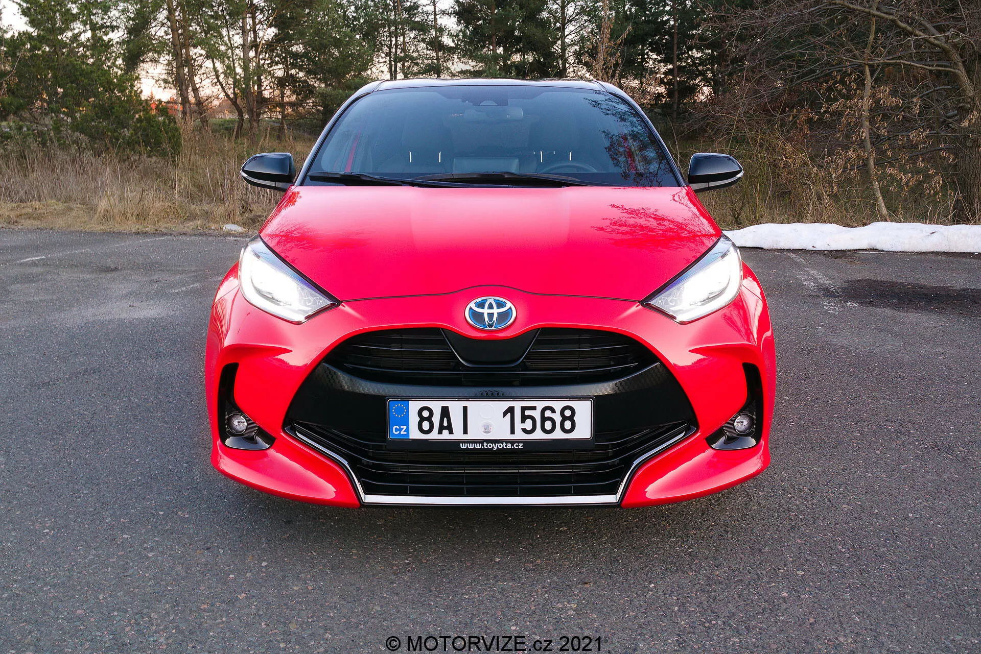 L'immagine mostra una vista frontale di una Toyota Yaris 2020 rossa, presa con un angolo leggermente inclinato e focalizzata sulla griglia frontale, sui fari e sull'area del cofano, con la targa completamente visibile. L'auto presenta elementi di design distintivi, tra cui l'emblema Toyota centrato sulla griglia, fari angolari affilati che si assottigliano verso la griglia e curve complesse sul cofano, che mostrano il suo design aerodinamico e sportivo. La foto, scattata durante il giorno prima del tramonto, suggerisce che l'auto è parcheggiata su una superficie piana in un ambiente che potrebbe essere la tarda autunno o l'inizio della primavera.