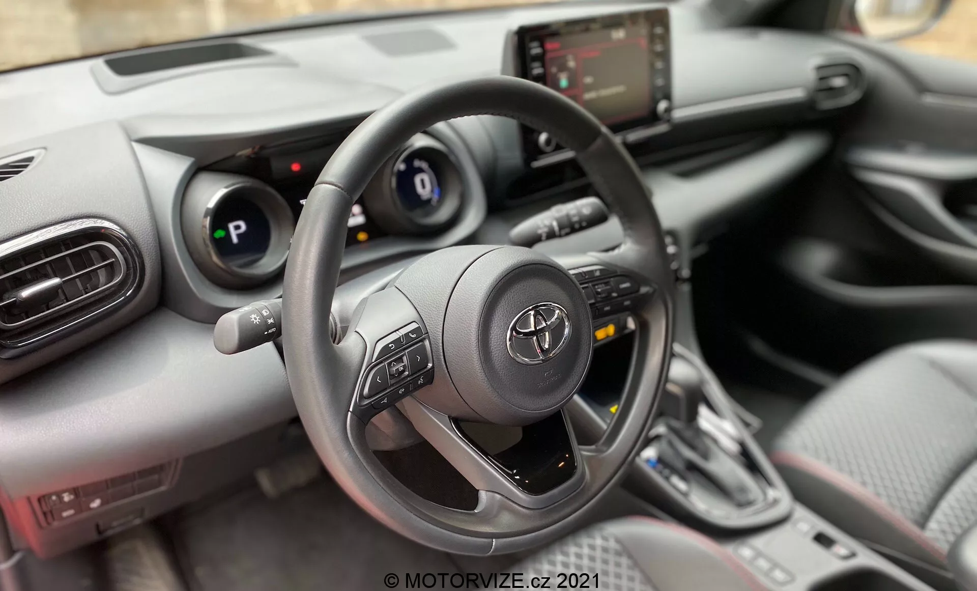 Questa immagine mostra l'interno di un'auto, concentrandosi sulla prospettiva del conducente. La ripresa è un primo piano, vista interna frontale, con l'angolazione della telecamera posizionata per catturare prevalentemente il volante, il cruscotto e la console centrale. La fotografia è probabilmente scattata dal sedile del conducente o possibilmente poco sopra, offrendo un punto di vista simile a quello di una persona seduta dietro il volante. Il volante è prominente in primo piano, centrato nell'inquadratura, e occupa una parte sostanziale dell'immagine. È di design a tre razze con comandi multifunzione montati sulle razze sinistra e destra. Dietro il volante, il quadro strumenti presenta un display digitale affiancato da indicatori analogici. A destra del volante, la console centrale include un touchscreen di infotainment, pannello di controllo del clima e selettore di marcia, indicando un veicolo moderno con funzioni interattive.