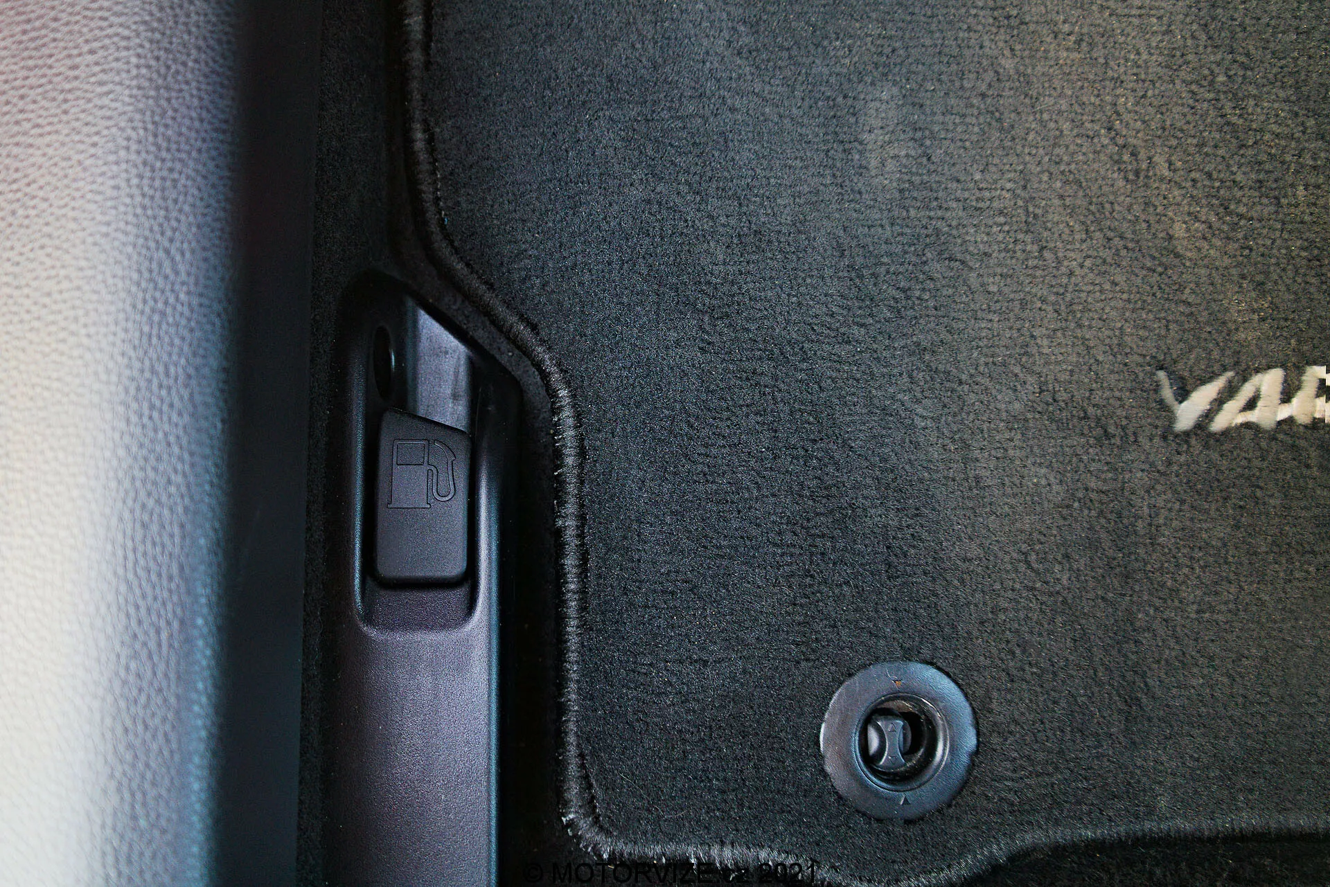 Vista ravvicinata dell'interno della Toyota Yaris 2020, catturando il lato inferiore sinistro vicino alla porta. Visibili un tappetino di colore scuro con testo ricamato parziale, un gancio di ritenzione circolare in metallo, il battitacco in plastica con rivestimento protettivo, una leva di apertura del bagagliaio che suggerisce il lato del conducente e uno schema a doppio tono con trame contrastanti di tappeto e plastica liscia.