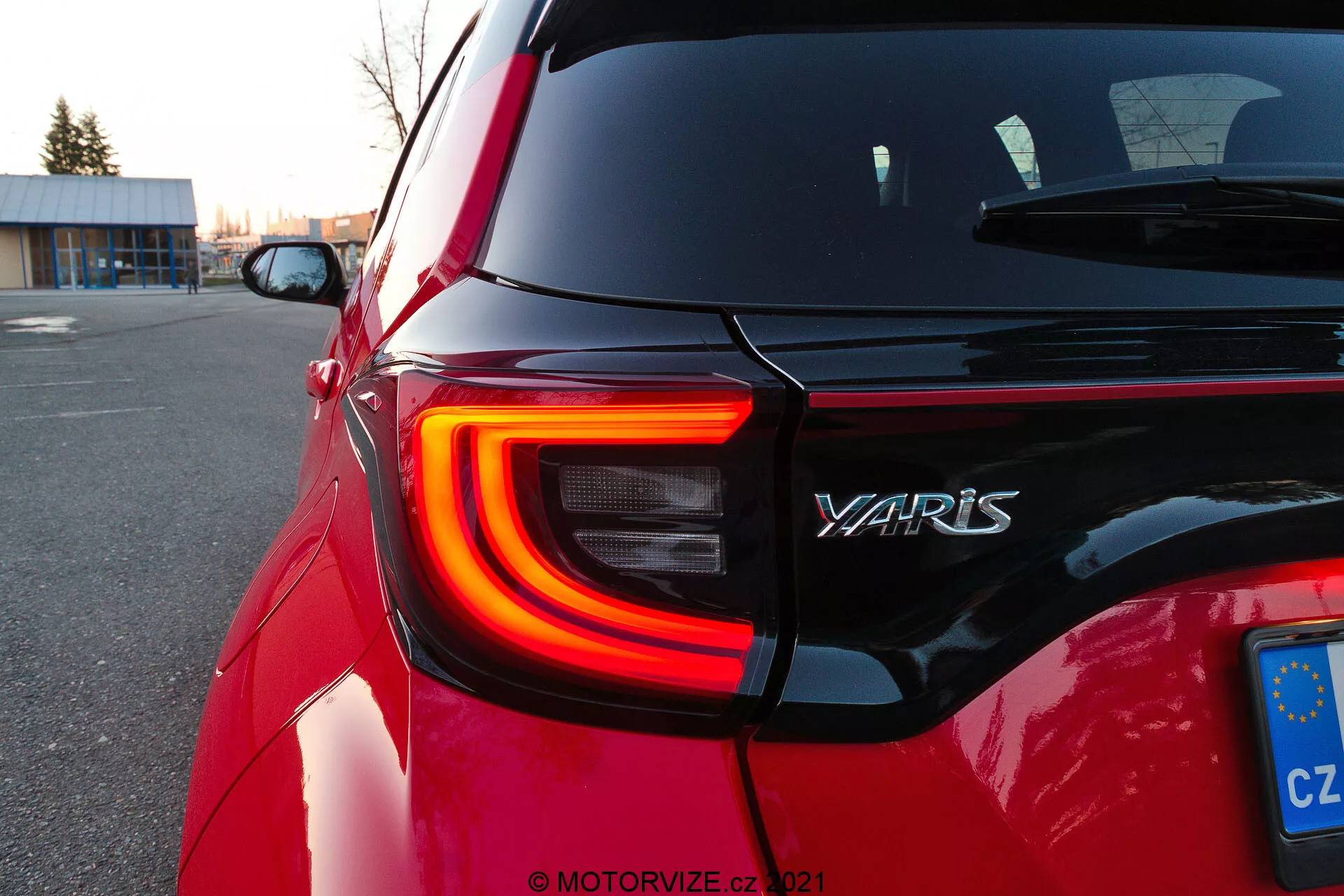 Primo piano della sezione posteriore di una Toyota Yaris rossa del 2020, che mostra la fanale posteriore sinistro a LED con la sua distintiva forma a C, il nome del modello 'YARIS' sul portellone e una targa dell'Unione Europea della Repubblica Ceca. L'angolo obliquo dello scatto mette in evidenza i contorni e la riflettività della vernice dell'auto al crepuscolo in un parcheggio, con sfondi sfocati che attirano l'attenzione sui dettagli del design.