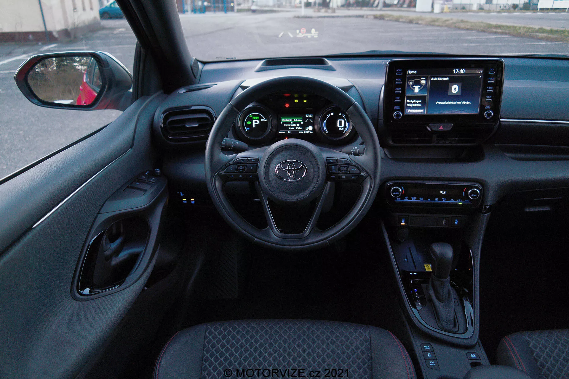 Prospettiva dell'interno dal sedile del conducente di una Toyota Yaris 2020, con volante con comandi integrati, quadro strumenti digitale e schermo per l'infotainment. Gli interni presentano pulsanti di controllo del clima, leva del cambio e una combinazione di colori prevalentemente nera con superfici testurizzate e accenti cromati.