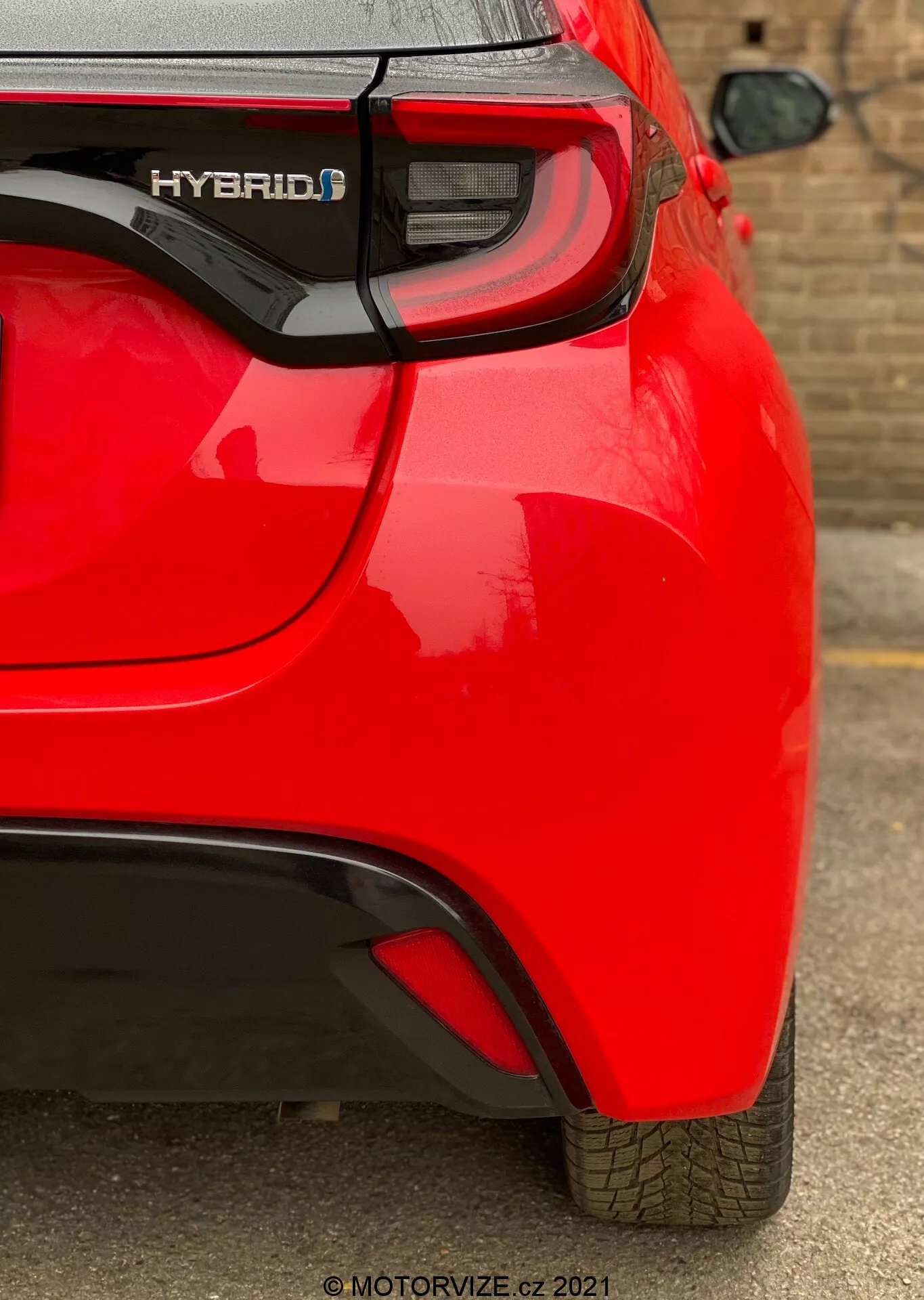 Vista posteriore angolata di una Toyota Yaris ibrida rossa del 2020, con focus sulla quartiera posteriore destra. Cattura caratteristiche come il badge 'HYBRID', il moderno gruppo ottico posteriore a LED, una leggera piegatura aerodinamica sul paraurti posteriore e un sensore di parcheggio visibile. Vernice lucida e battistrada ben mantenuto indicano un veicolo nuovo o ben curato.