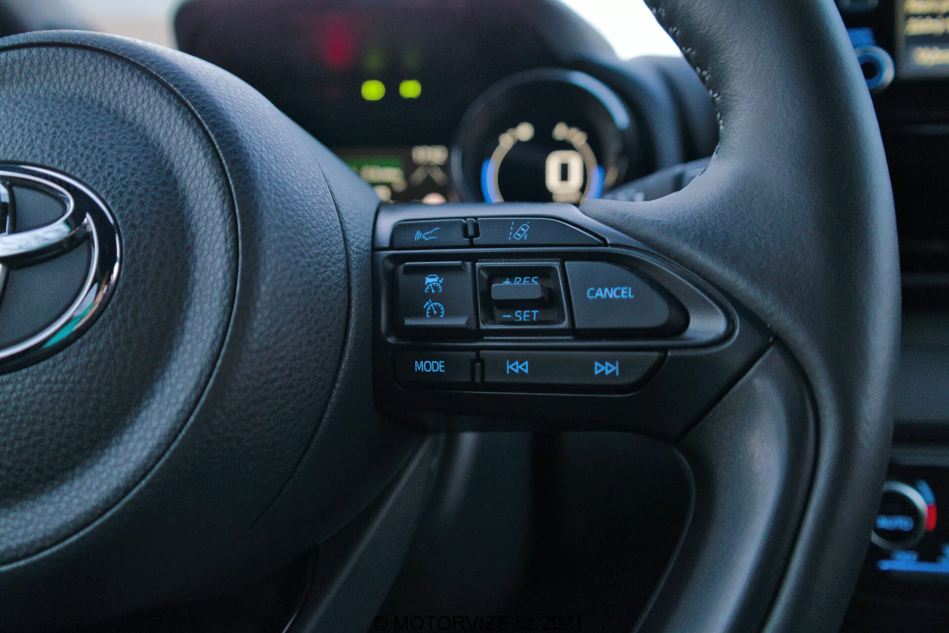 Immagine degli interni di una Toyota Yaris 2020 dal sedile del conducente, focalizzata sul lato sinistro del volante multifunzione. I pulsanti tattili, inclusi l'icona della funzione di sicurezza, controllo di crociera con etichetta 'SET', pulsante di annullamento, 'MODE' per multimedia e controlli del volume '+', '-', sono chiaramente visibili. Il veicolo è in modalità 'P' parcheggio, con il quadro strumenti e gli elementi di sfondo leggermente sfocati.