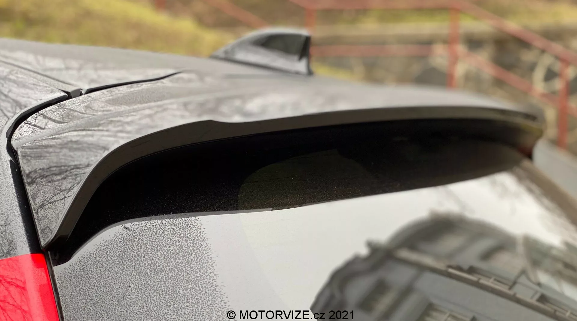 Primo piano dello spoiler posteriore della Toyota Yaris 2020 con vernice nera in contrasto con la carrozzeria metallica. L'immagine cattura gocce d'acqua sullo spoiler, suggerendo condizioni di bagnato, e un profilo laterale che rivela il suo design leggermente elevato per una maggiore aerodinamica. Lo sfondo sfocato all'aperto aggiunge contesto alla finitura premium dell'auto e la luce posteriore LED visibile suggerisce caratteristiche moderne.