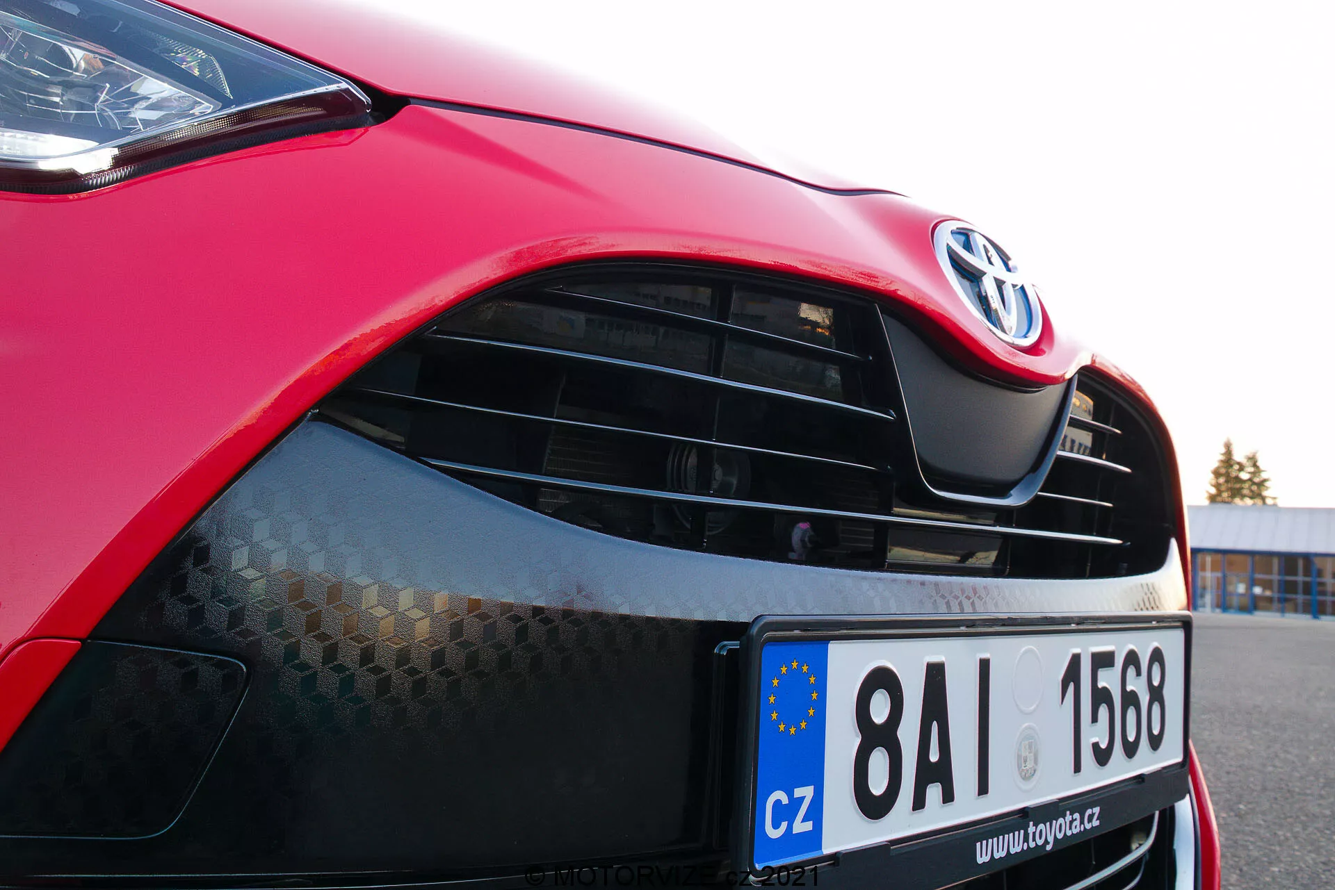 Primo piano di una Toyota Yaris 2020 rossa focalizzato sul lato anteriore sinistro, evidenziando il cofano, il moderno faro con potenziale tecnologia LED o HID, la griglia con emblema Toyota e motivo a nido d'ape, vernice rossa lucida e paraurti anteriore parzialmente visibile con presa d'aria inferiore. La targa della Unione Europea indica registrazione nella Repubblica Ceca.