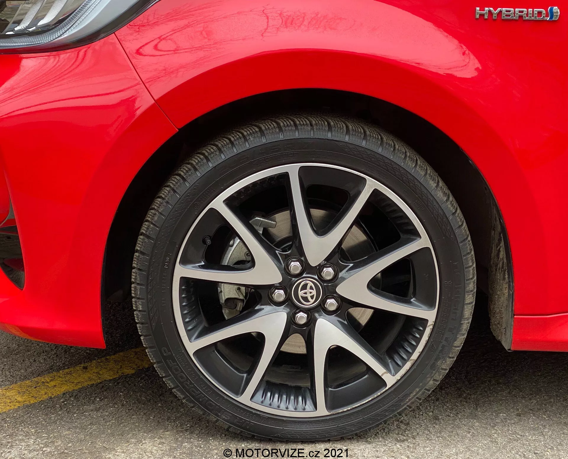 Vista ravvicinata della ruota anteriore e parte del lato di una Toyota Yaris 2020 ibrida rossa, presa dal livello del suolo. Il cerchio in lega a più razze presenta una finitura argento e nero. Il battistrada del pneumatico è chiaramente visibile e non mostra segni di usura. La spalla del pneumatico include specifiche stampate e il badge 'HYBRID' è chiaramente visibile sopra la ruota sul lato della macchina. L'illuminazione diurna uniforme migliora la visibilità del design della ruota e del marchio ibrido.