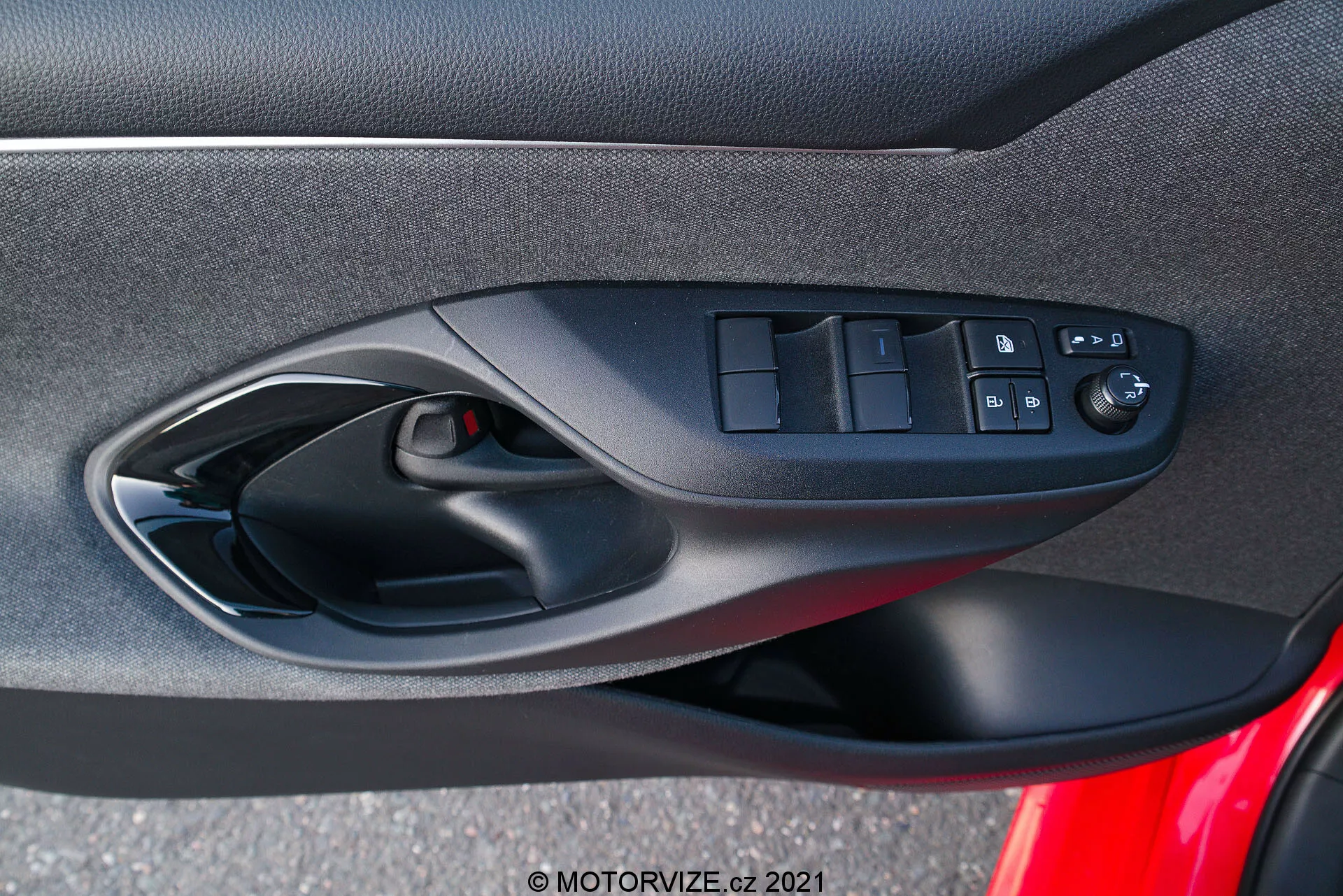 Pannello interno della portiera di una Toyota Yaris 2020 che si concentra sui controlli ergonomicamente progettati della portiera lato conducente, inclusi i comandi dei finestrini elettrici, il pulsante di blocco delle portiere e i controlli regolabili elettricamente degli specchietti, catturato dalla prospettiva di un occupante all'interno del veicolo. La foto mette in evidenza la combinazione di materiali in tessuto morbido o composito, lo schema di colore nero e una griglia dell'altoparlante visibile come parte del sistema audio dell'auto.