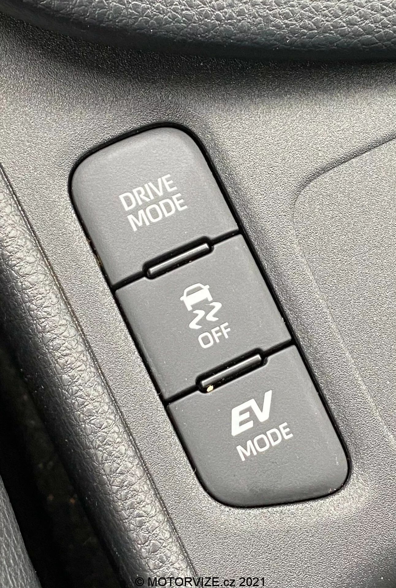 Scatto ravvicinato dell'interno di una Toyota Yaris 2020, focalizzato su tre pulsanti di controllo allineati verticalmente relativi alle modalità di guida. Dall'alto verso il basso: 'DRIVE MODE' con un'icona di strada tortuosa, 'Traction Control OFF' con un'icona di auto che slitta e 'EV MODE', che indica la funzione veicolo elettrico. I pulsanti sono montati su una superficie testurizzata, probabilmente parte di un pannello della portiera o della consolle centrale. L'immagine è nitidamente a fuoco sui pulsanti, con le texture circostanti leggermente sfocate, illuminata uniformemente dalla luce del giorno per una chiara visibilità.
