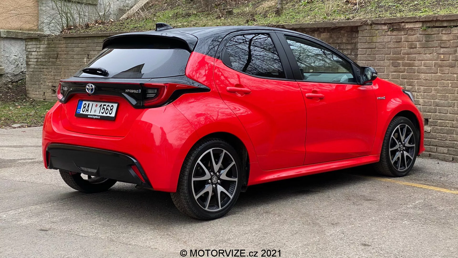 Vista di tre quarti posteriore di un'auto hatchback moderna di colore rosso brillante, con linee e pieghe nitide, spoiler sul tetto, luci posteriori a LED, antenna a pinna di squalo, cerchi in lega multi-razziali e posizionata in un parcheggio con vernice riflettente e ben curata, suggerendo un modello recente o ben tenuto in una versione sportiva.