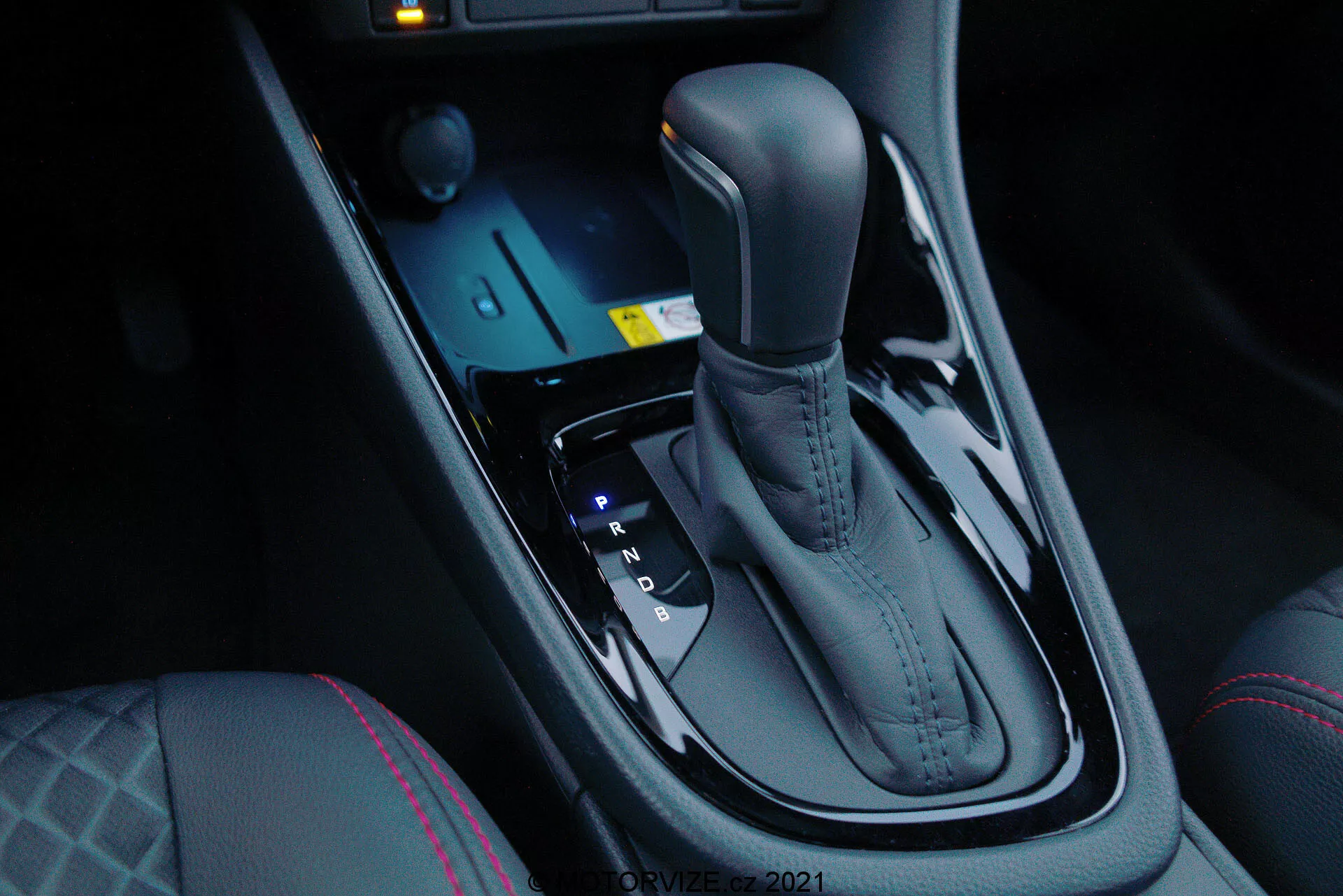 Vista ravvicinata della leva del cambio automatico della Toyota Yaris 2020, vista dal lato del passeggero anteriore ad un angolo verso il basso, che mette in risalto la finitura metallica elegante e il design ergonomico. La leva del cambio è posizionata al centro tra i sedili del conducente e del passeggero anteriore e include posizioni Park, Reverse, Neutral e Drive, nonché opzioni di cambio manuale (+ e -). Davanti alla leva del cambio ci sono controlli elettronici per le modalità di guida e il controllo della trazione, insieme a un sistema di avviamento a pulsante illuminato di blu. La consolle centrale scura e lucida aggiunge un tocco di lusso moderno, completato dai comandi del riscaldamento dei sedili e dalle cuciture distintive sul sedile del conducente, indicando un comfort maggiore e dinamiche di guida avanzate.