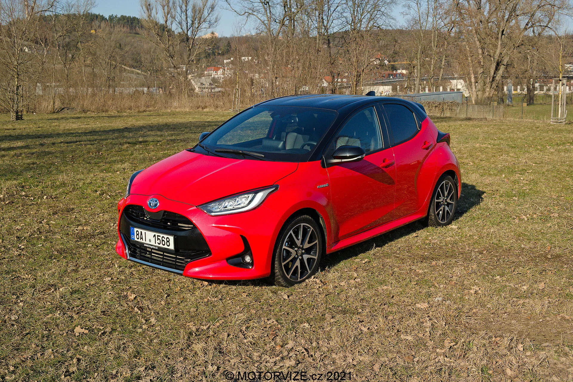L'immagine mostra una vibrante Toyota Yaris rossa del 2020, su un terreno erboso irregolare in un'area semi-rurale, vista da una prospettiva a tre quarti. Il design moderno e aerodinamico dell'auto presenta angoli acuti intorno ai fari e un aspetto compatto e sportivo. Le ruote a più razze scure e i fari LED stilizzati sono visibili. I finestrini laterali e il parabrezza anteriore offrono uno sguardo oscuro all'interno, suggerendo possibili sistemi avanzati di automazione.