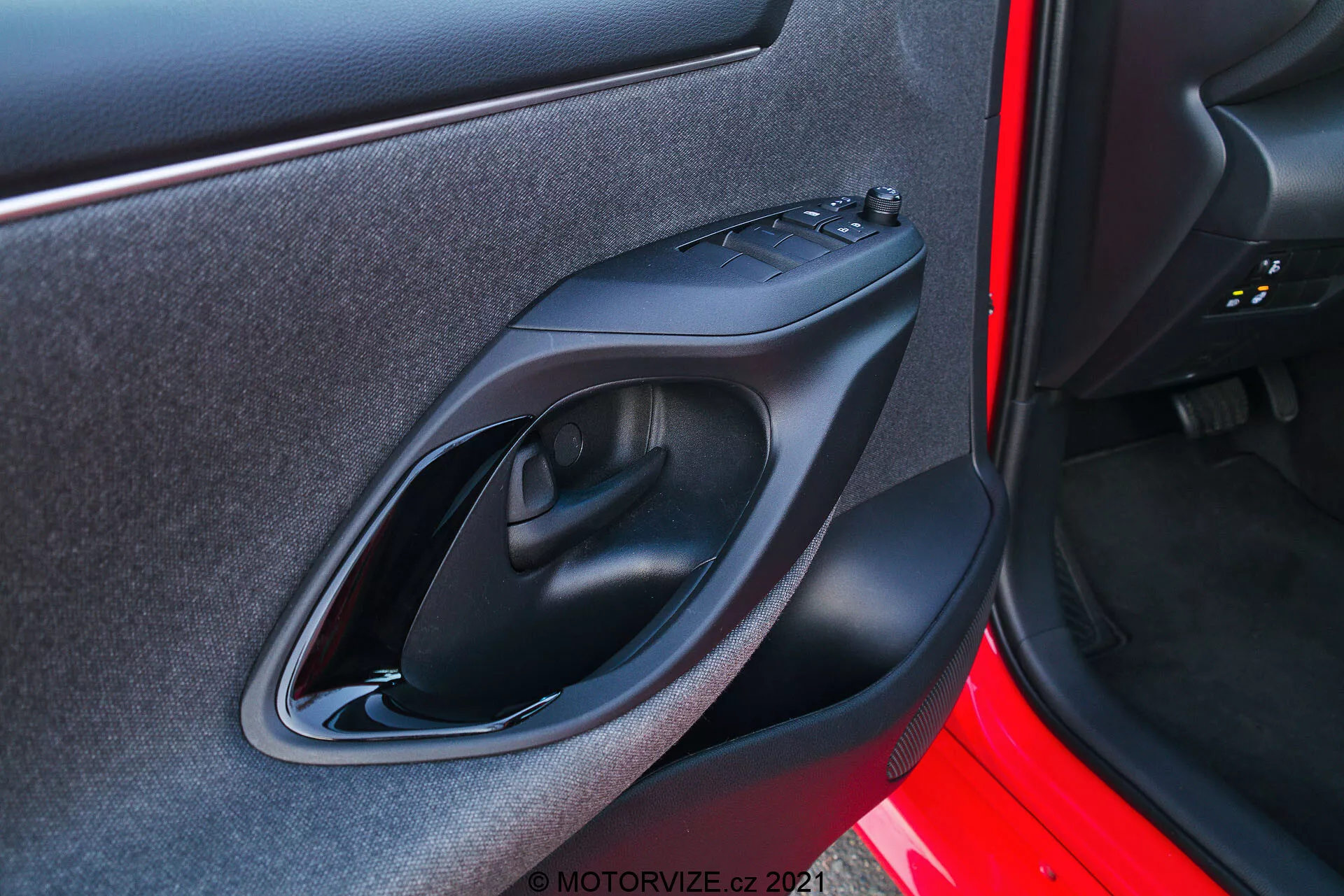 Primo piano del pannello interno della porta di una Toyota Yaris 2020 ripreso da una prospettiva esterna. L'immagine si concentra sul pannello della porta del lato destro con caratteristiche di design e tecnologiche come maniglia della porta in plastica nera opaca, pannello di controllo dei finestrini elettrici, bracciolo ergonomico sagomato e una piccola tasca di stoccaggio. L'esterno della porta è verniciato in un colore rosso vibrante.