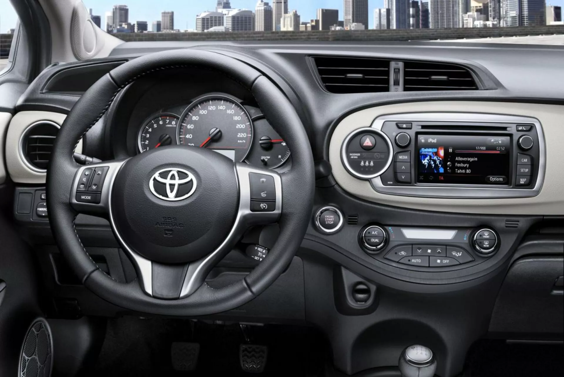 Vista interna della Toyota Yaris 2014 dalla prospettiva del conducente che evidenzia il volante a tre razze con emblema Toyota, comandi montati per audio e telefono vivavoce, tachimetro e contagiri analogici con un display digitale, e un sistema di infotainment moderno con pulsanti per il controllo del clima sul cruscotto