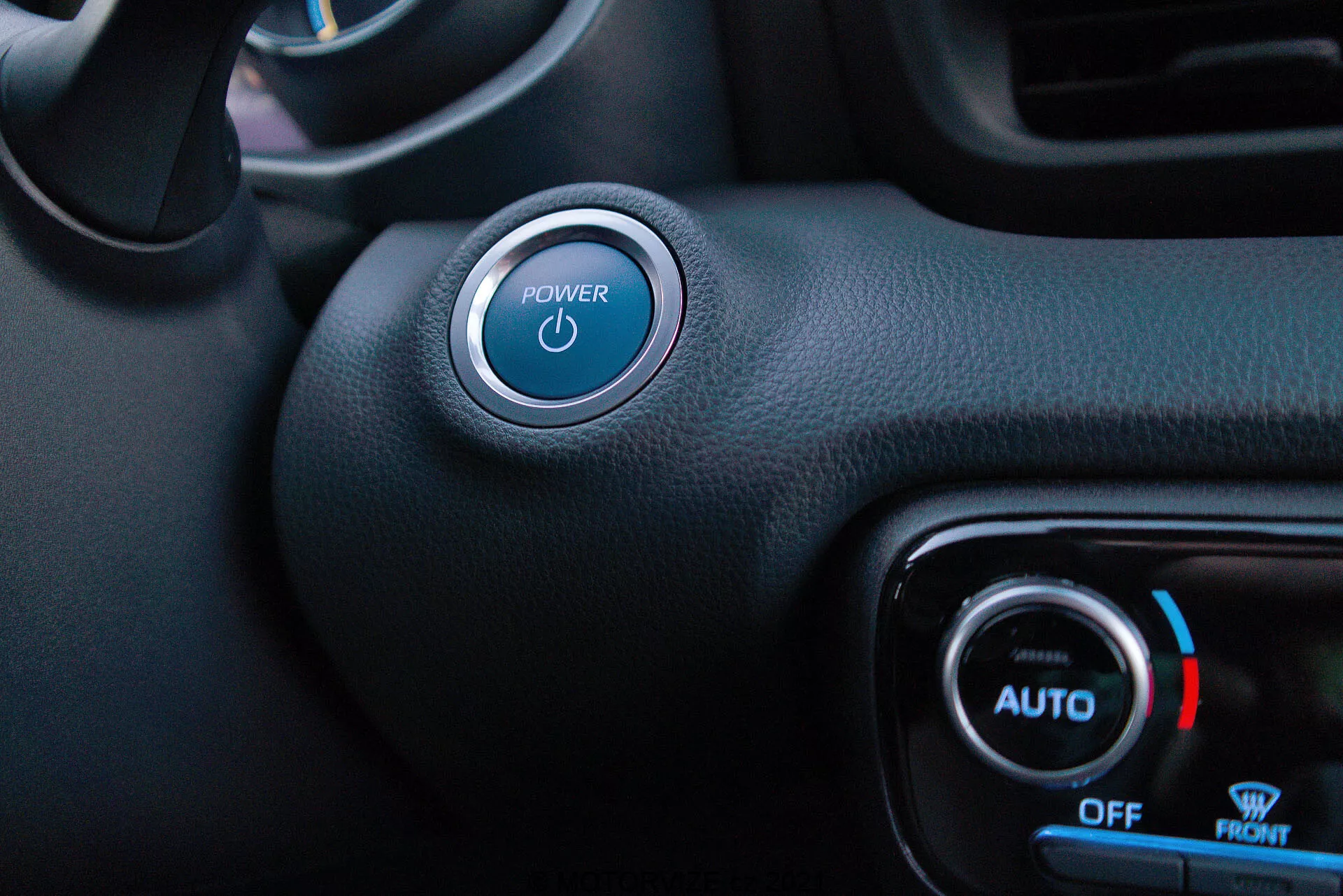 Vista degli interni dal lato del conducente della Toyota Yaris 2020 con evidenza del pulsante Start/Stop e del controllo del clima. Il pulsante di accensione con simbolo illuminato blu è prominente in alto a sinistra, con un accento argento. In basso a destra, le manopole del controllo del clima mostrano una luce blu e un'icona fiocco di neve per la temperatura fredda, con etichette 'AUTO', 'OFF' e 'FRONT'. Una combinazione di materiali sintetici neri testurizzati e lisci denota uno stile minimalista con funzioni di avvio senza chiave e controllo automatico del clima.